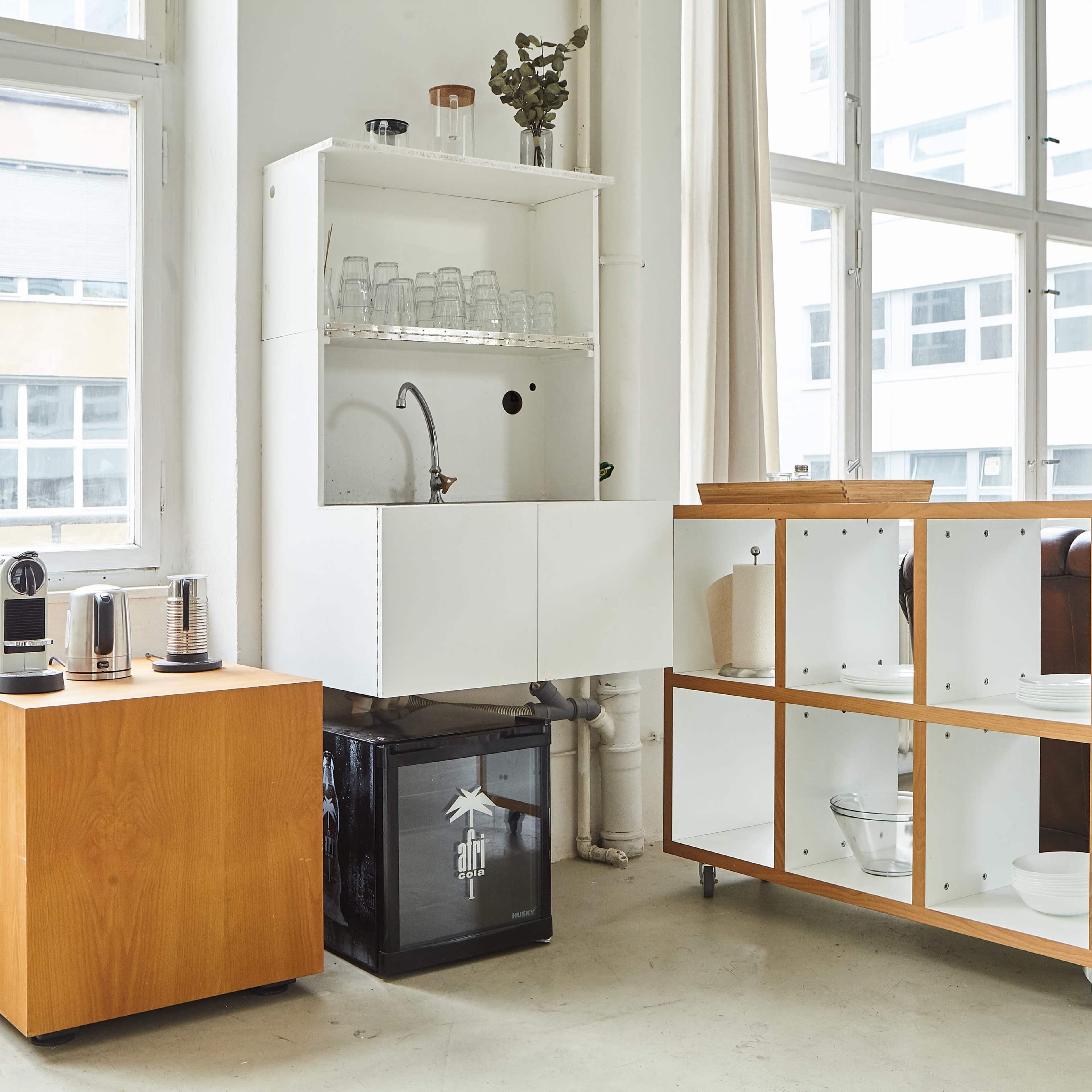 A modern kitchen space featuring a wooden cabinet with an integrated sink, a countertop espresso machine, and a modular shelving unit wheel on casters.