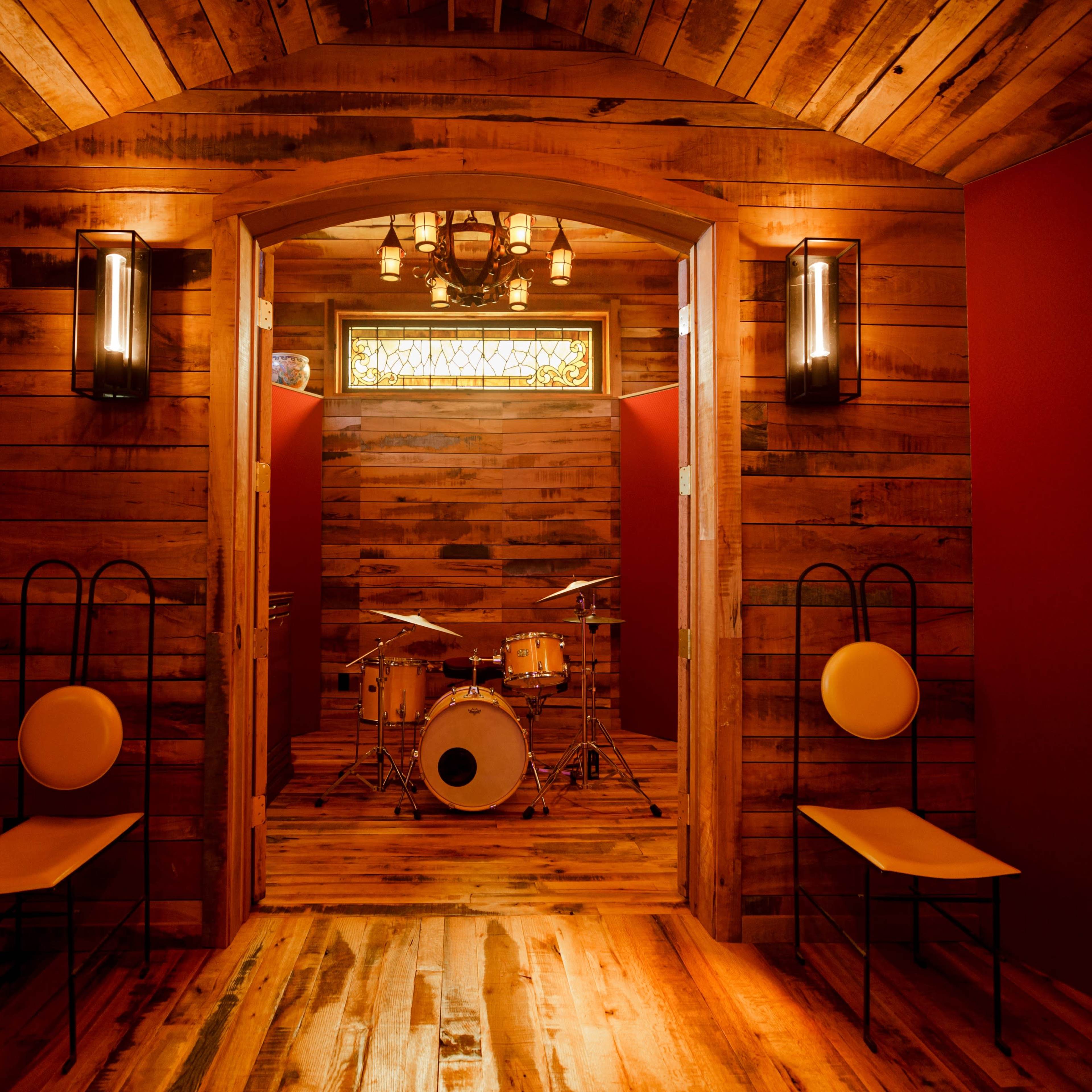 A wooden room features two chairs and a view of a drum set in the background, illuminated by warm lighting.
