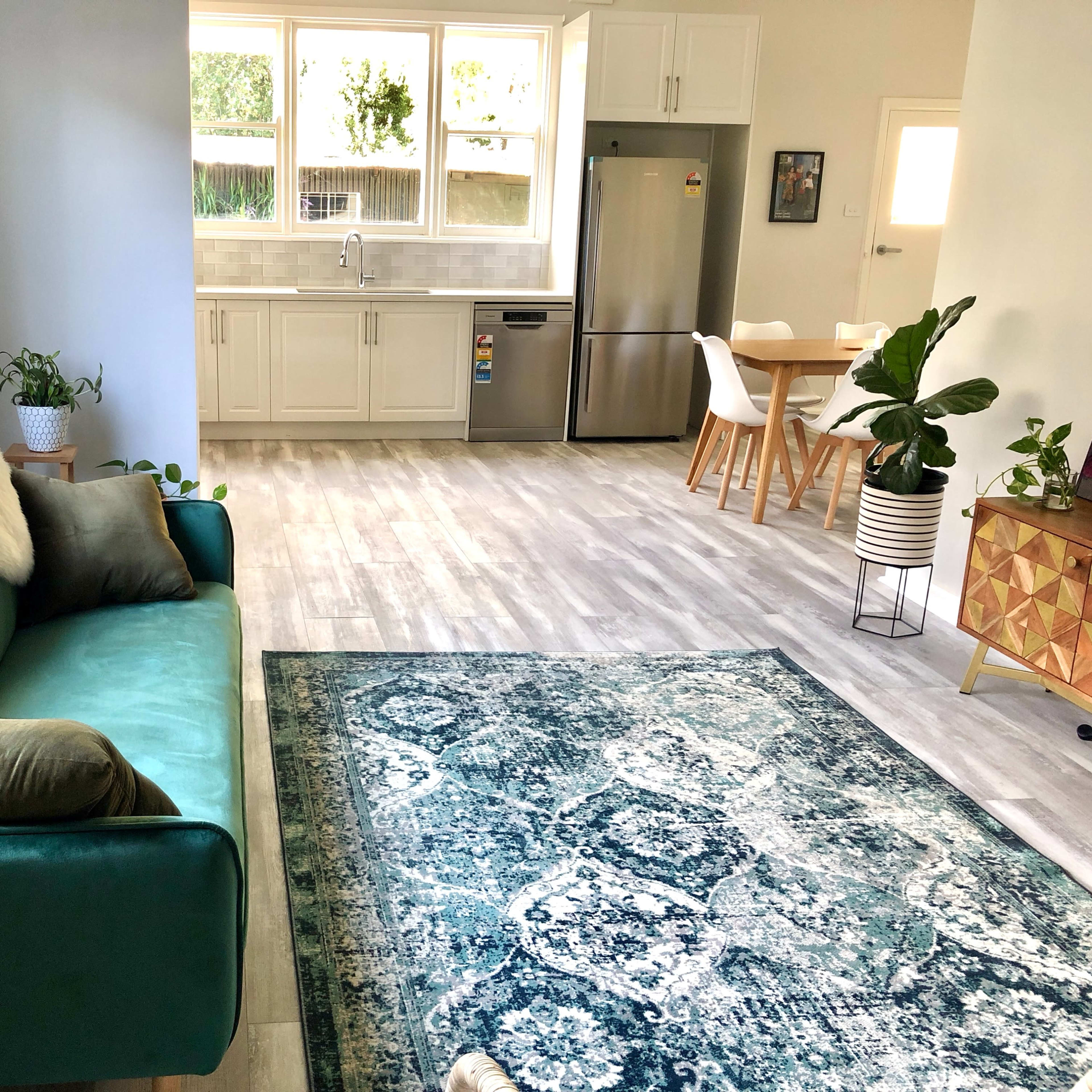 A modern living space with a green sofa, a patterned blue rug, a dining area with a wooden table and chairs, and a kitchen featuring white cabinetry and stainless steel appliances.