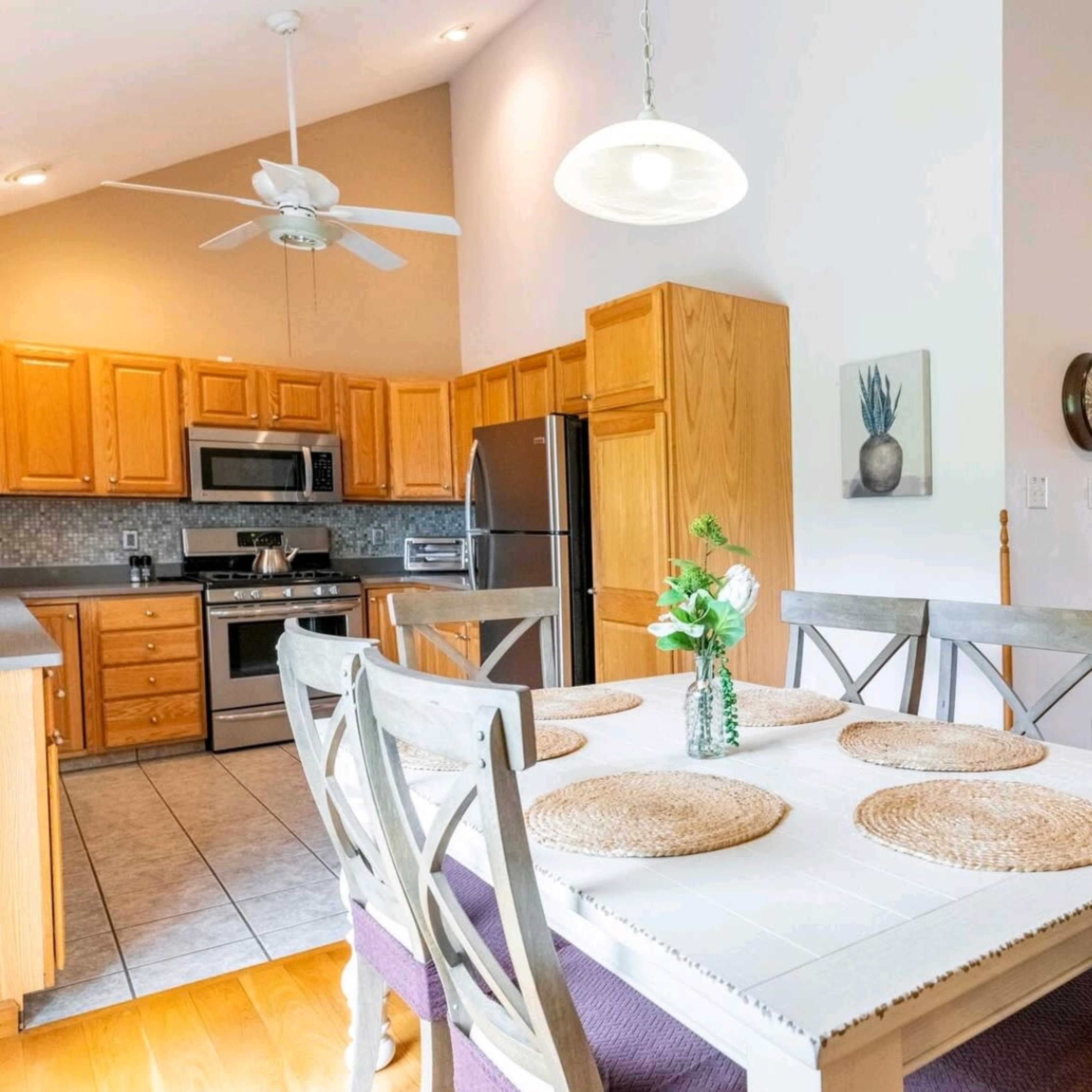 A dining area features a wooden table set with placemats, surrounded by chairs, and is adjacent to a kitchen with light wood cabinets and stainless steel appliances.