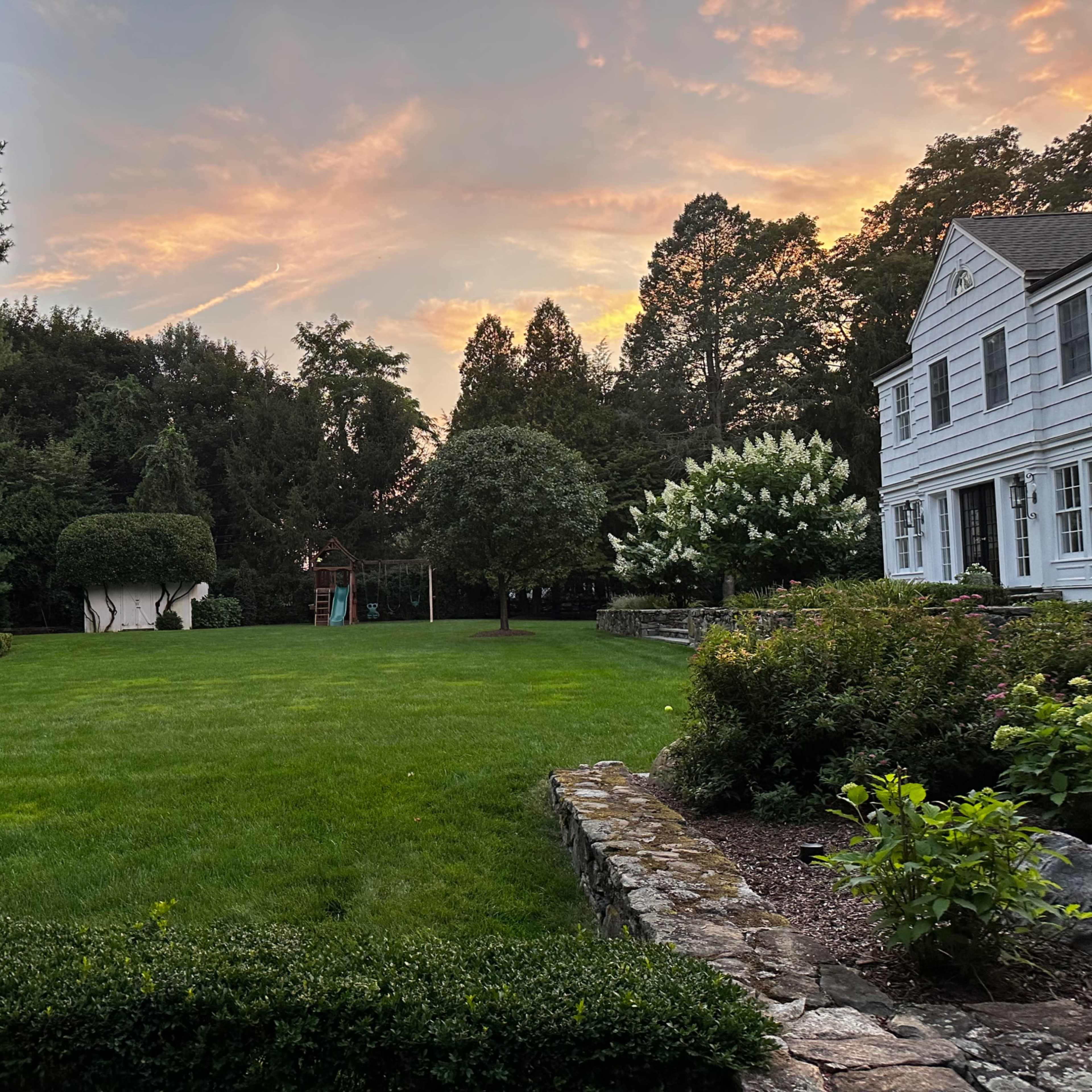 A well-manicured lawn in a residential yard, bordered by trees and shrubs, with a house visible on the right and a colorful sunset in the background.