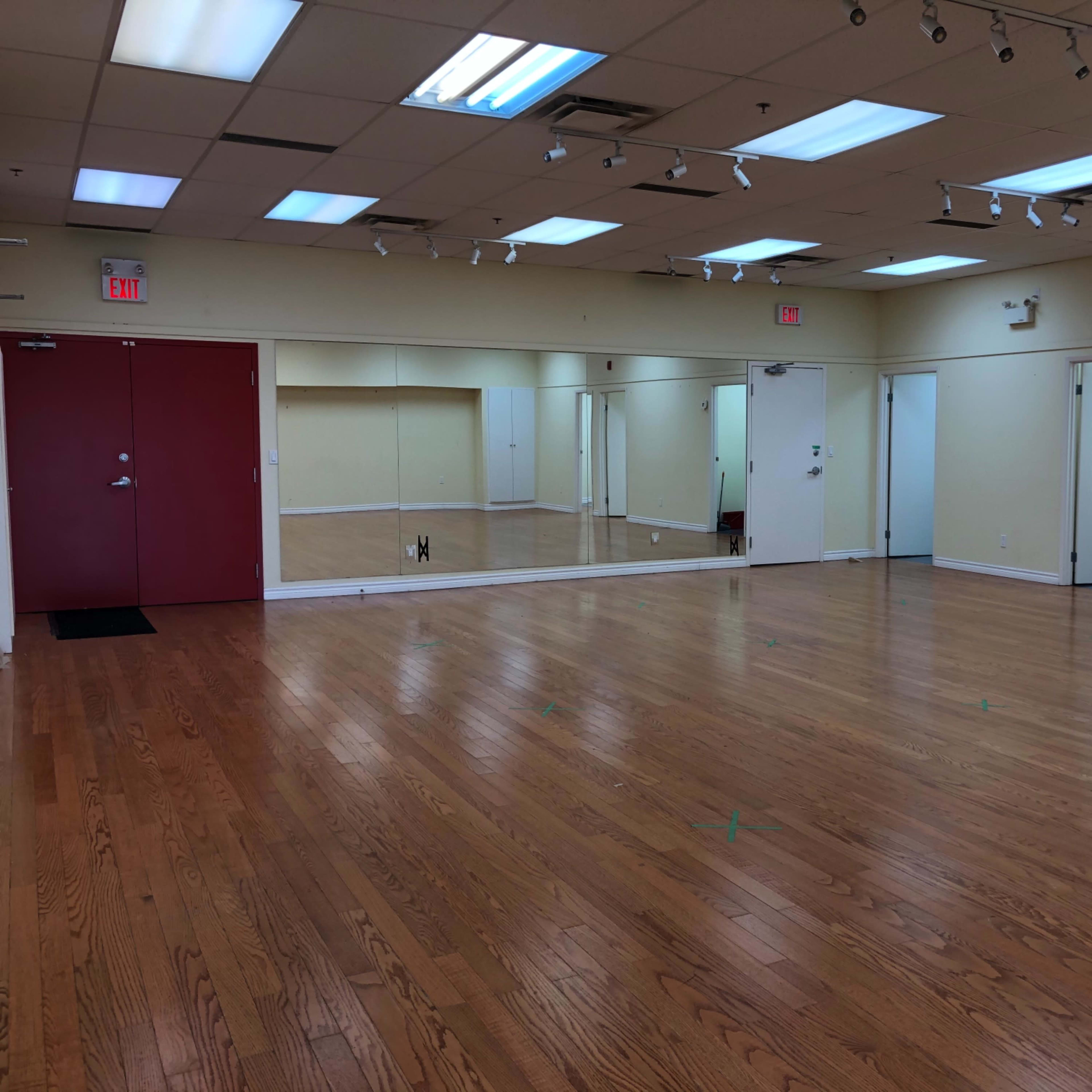 A spacious, empty room with hardwood floors, bright overhead lights, and several mirrors on one wall.