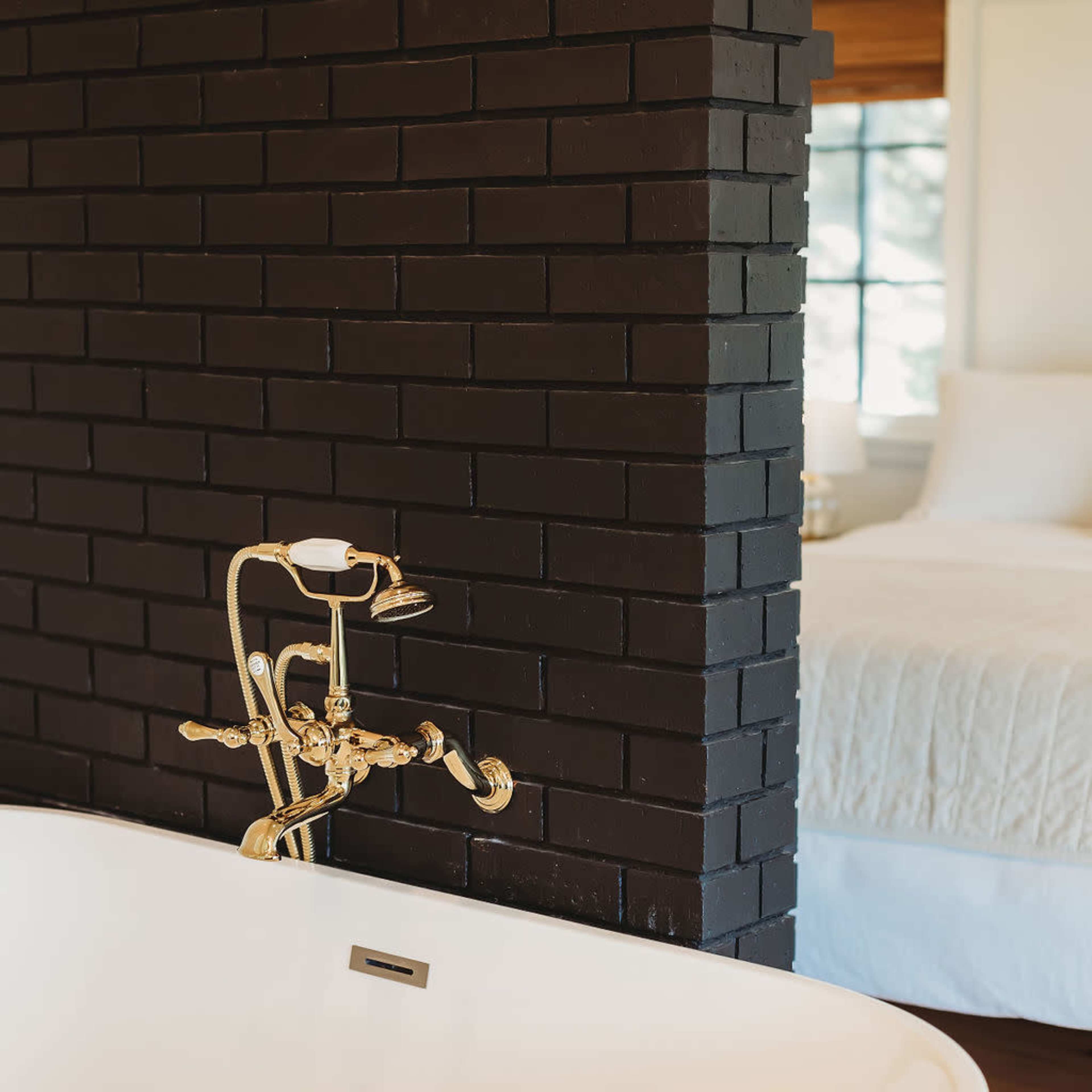 A freestanding white bathtub with a gold faucet is positioned in front of a black brick wall, with a glimpse of a bedroom in the background.