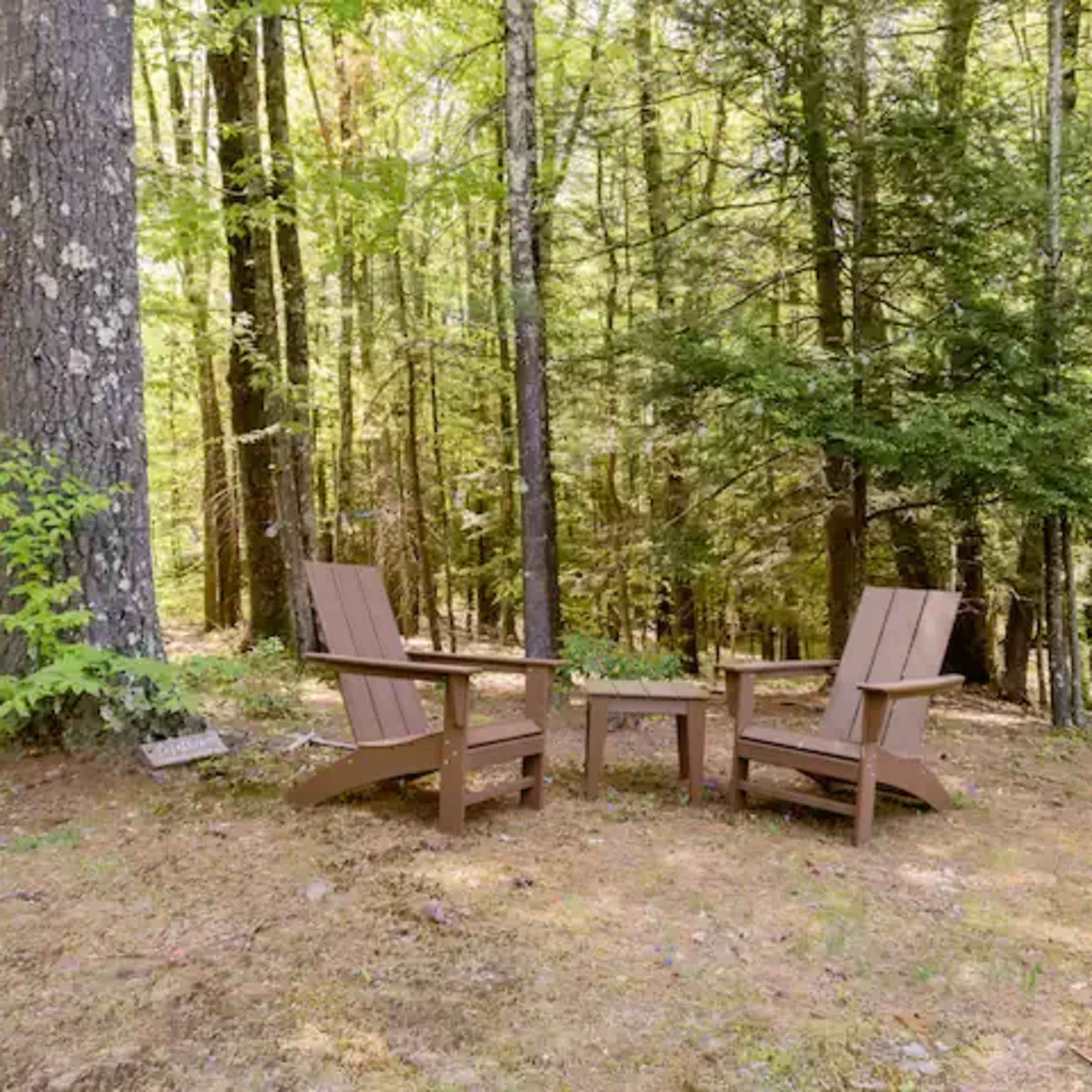 Two wooden Adirondack chairs and a small table are set in a forest clearing surrounded by trees.