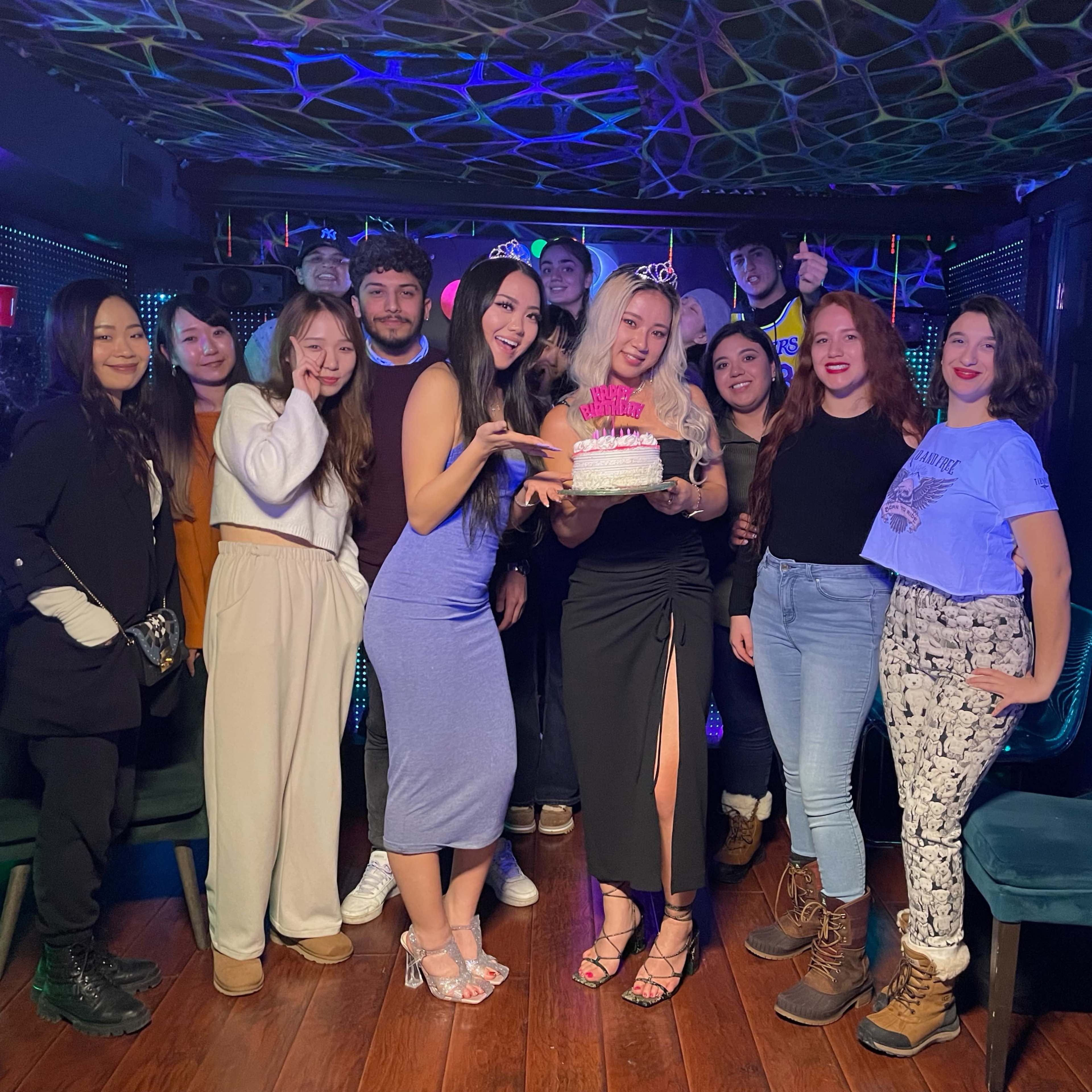 A group of twelve people gathers in a decorated interior space, smiling around a birthday cake.