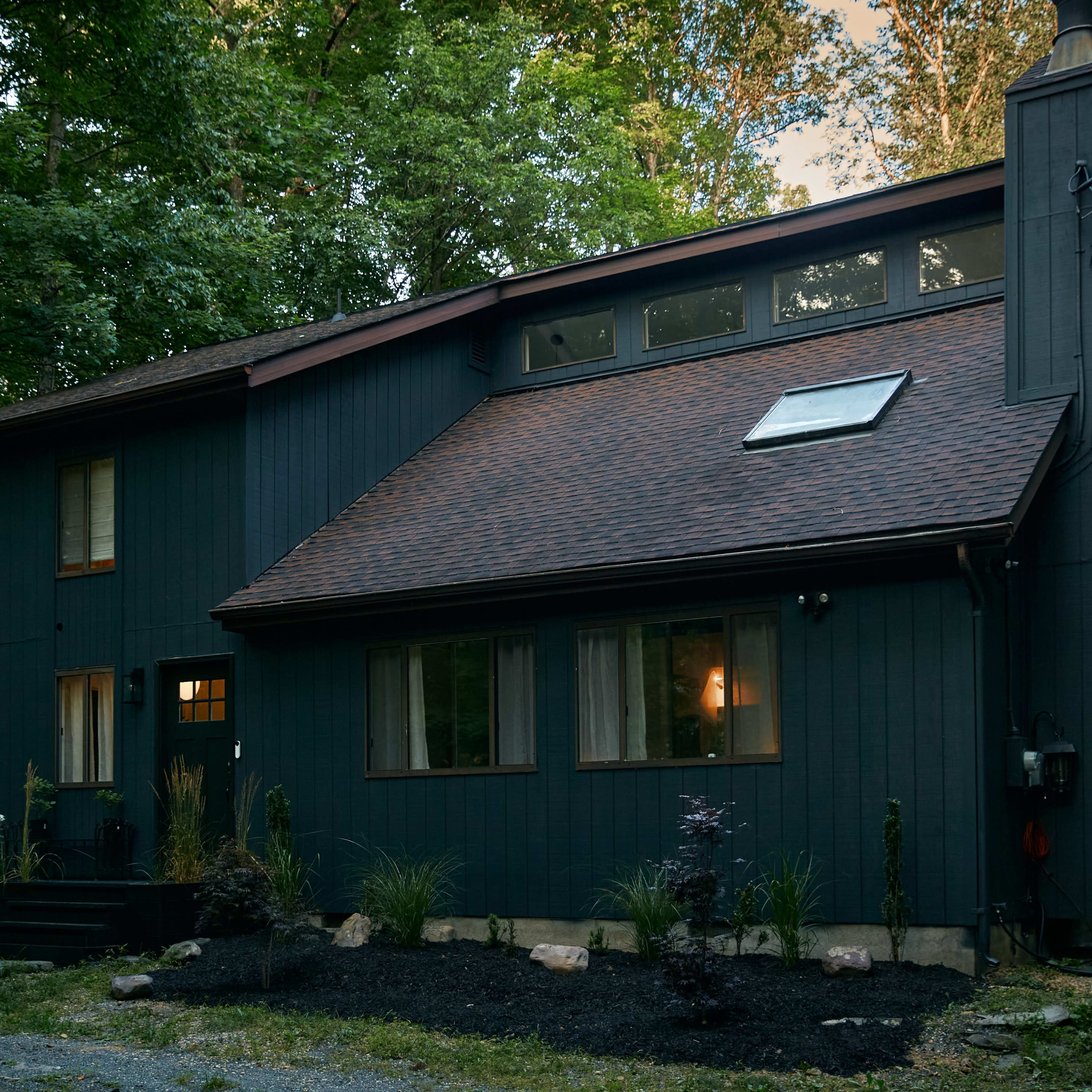A two-story dark blue house with a sloped roof is surrounded by trees and features a landscaped garden with rocks and plants.