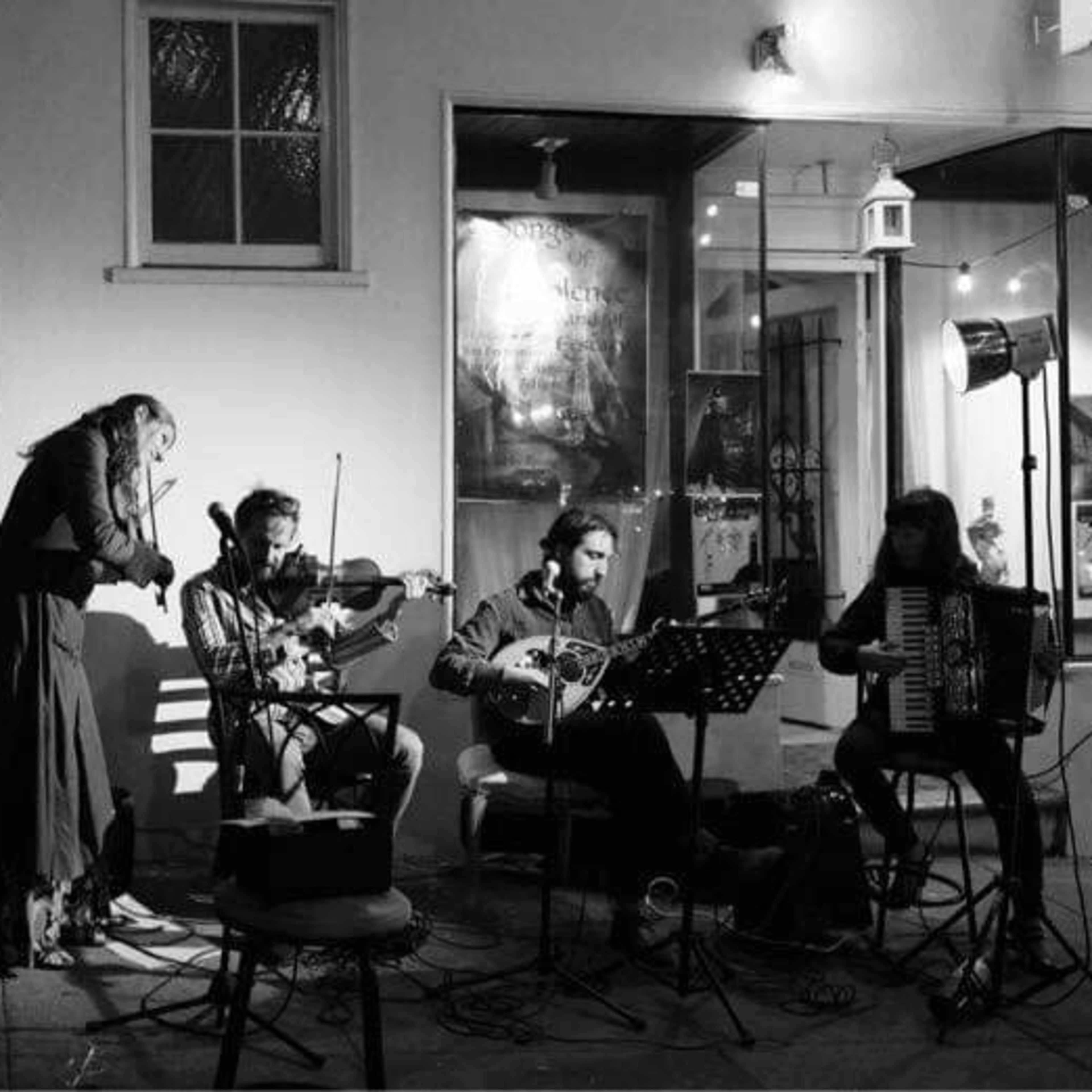 A group of five musicians performs in a dimly lit outdoor setting, with various instruments including a violin, accordion, and a stringed instrument, while one musician plays a hand drum.
