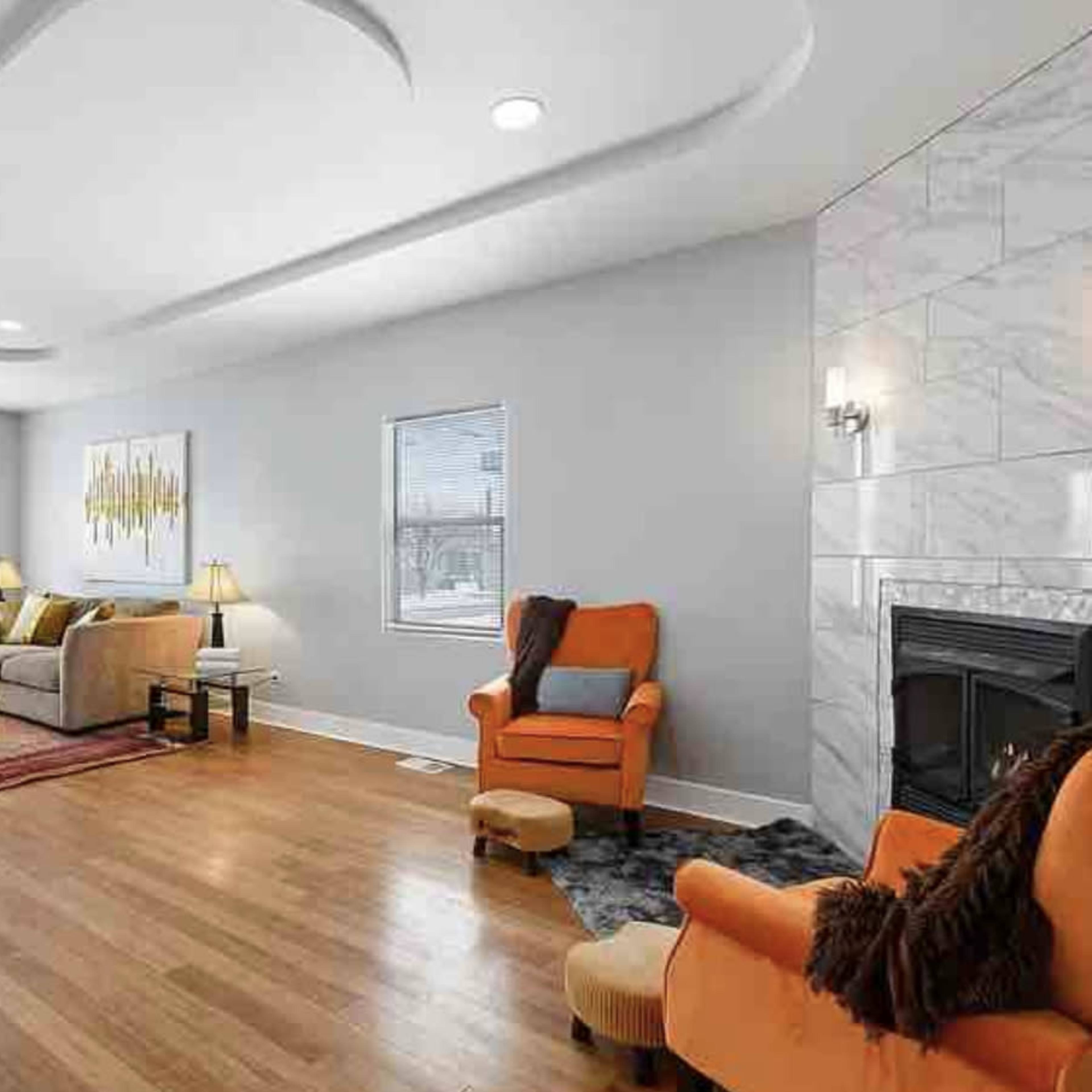 The living room features two gray sofas and an orange armchair arranged around a central rug, with a fireplace and large windows.