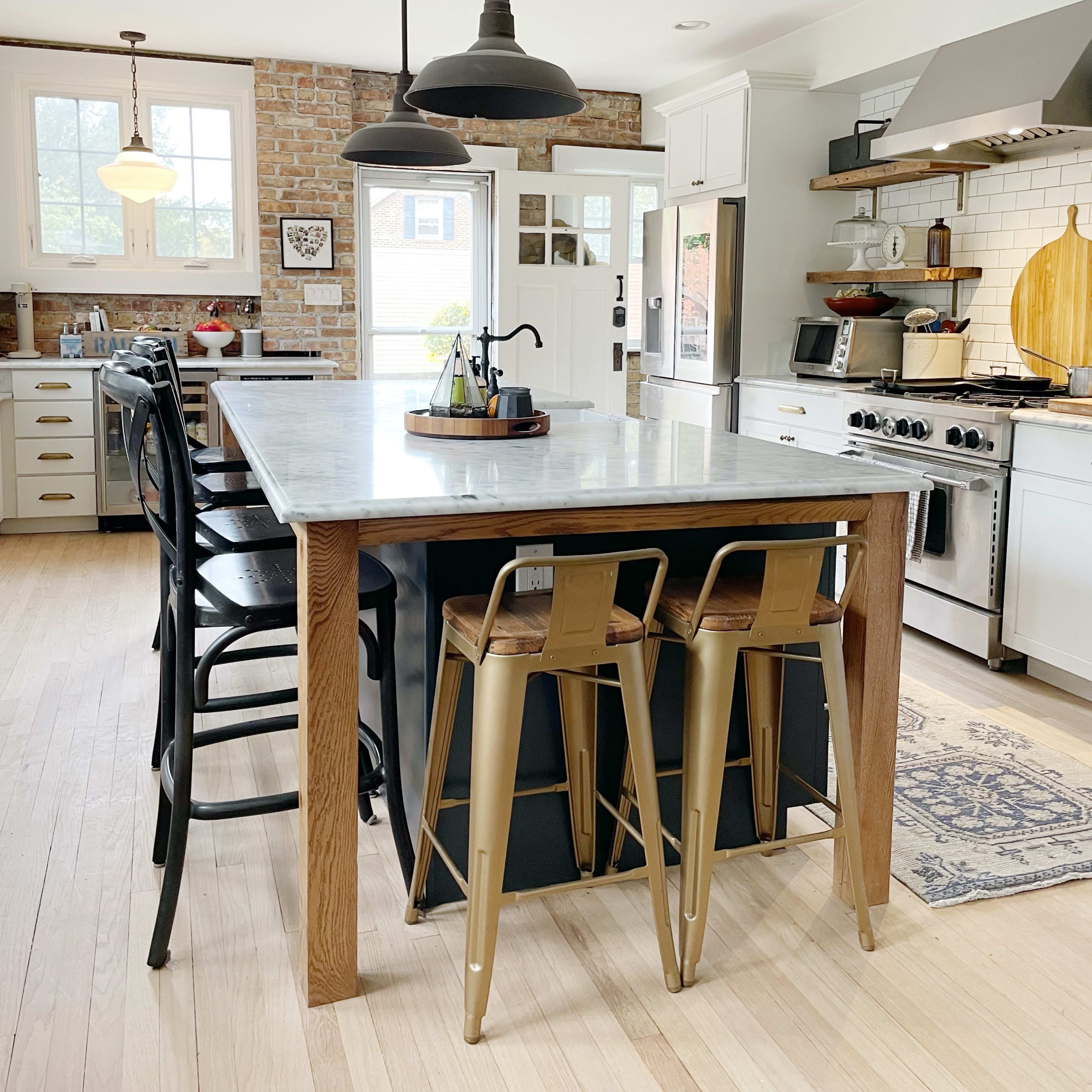 The kitchen features a large island with a marble countertop, black and gold bar stools, and stainless steel appliances, set against exposed brick and white cabinetry.