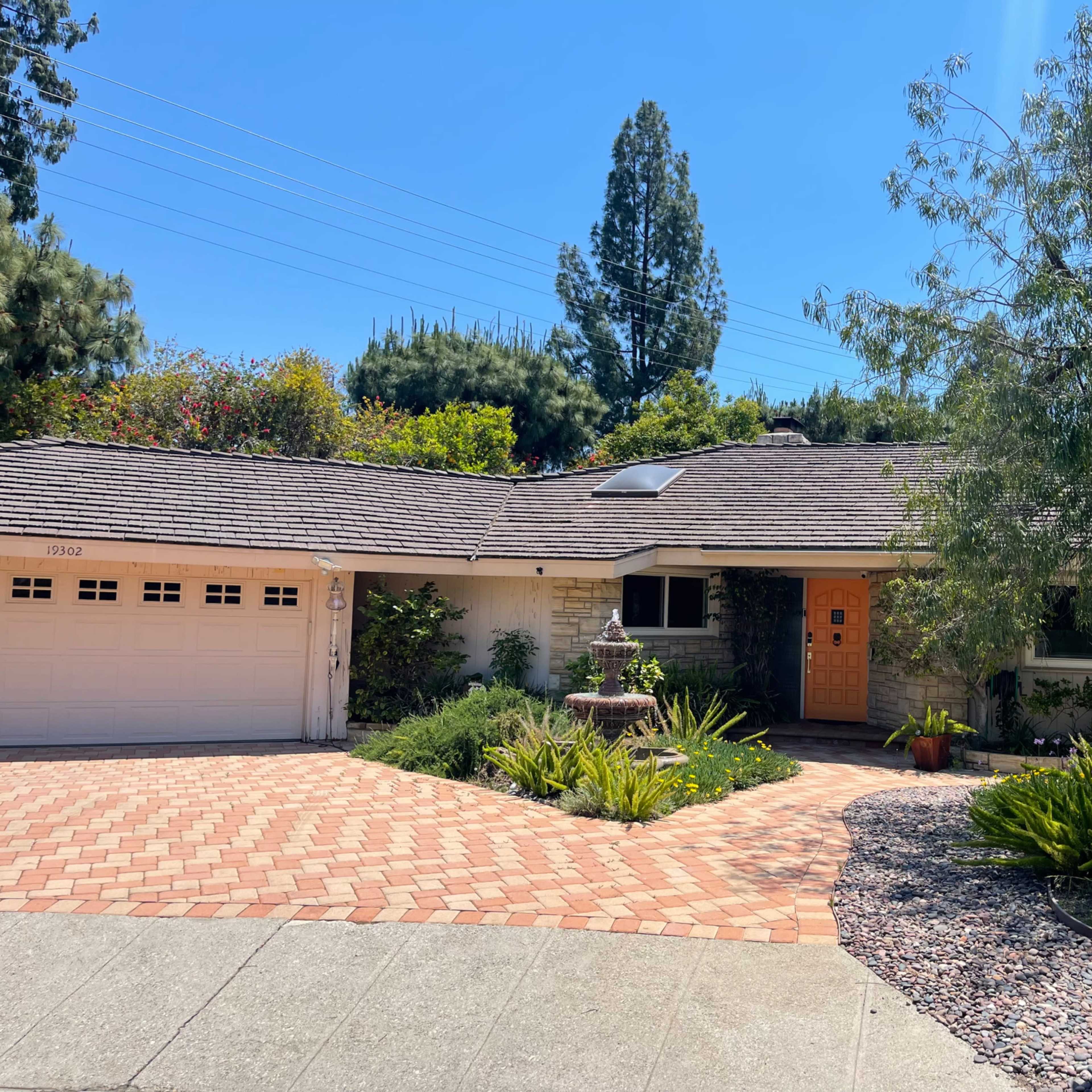 A single-story house with a brick driveway, surrounded by green plants and trees, features a garage on the left and a wooden front door.