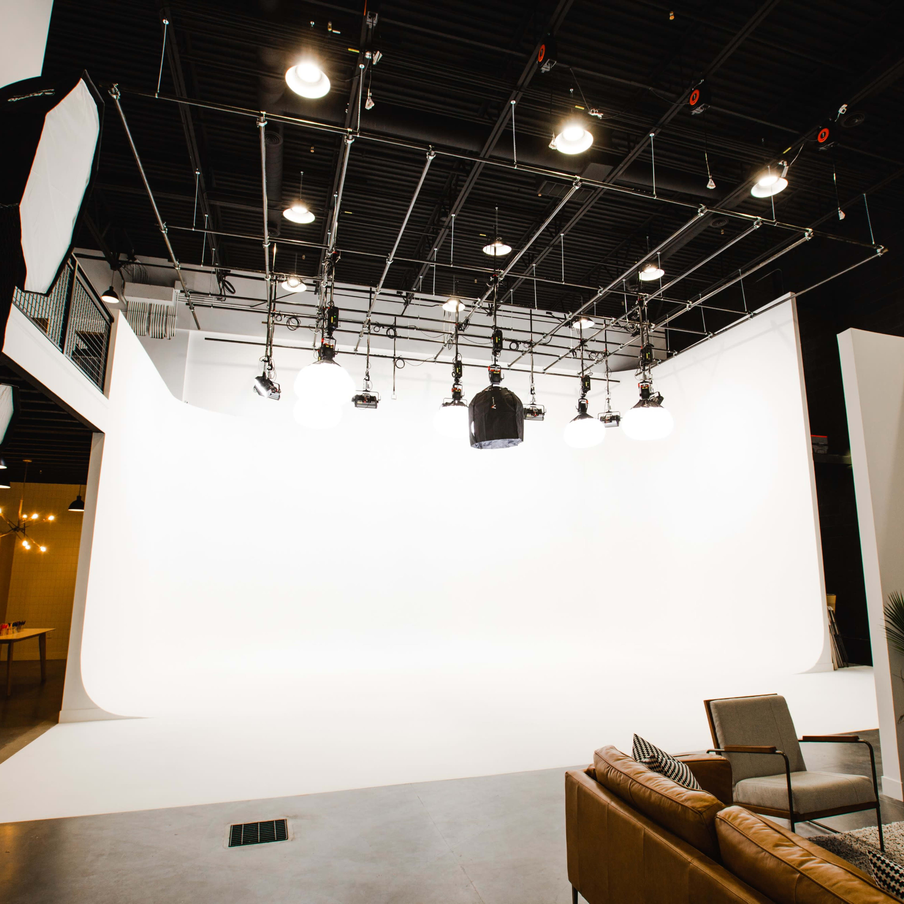 A modern studio space with a white backdrop, overhead lighting equipment, and a seating area with a couch and chairs.