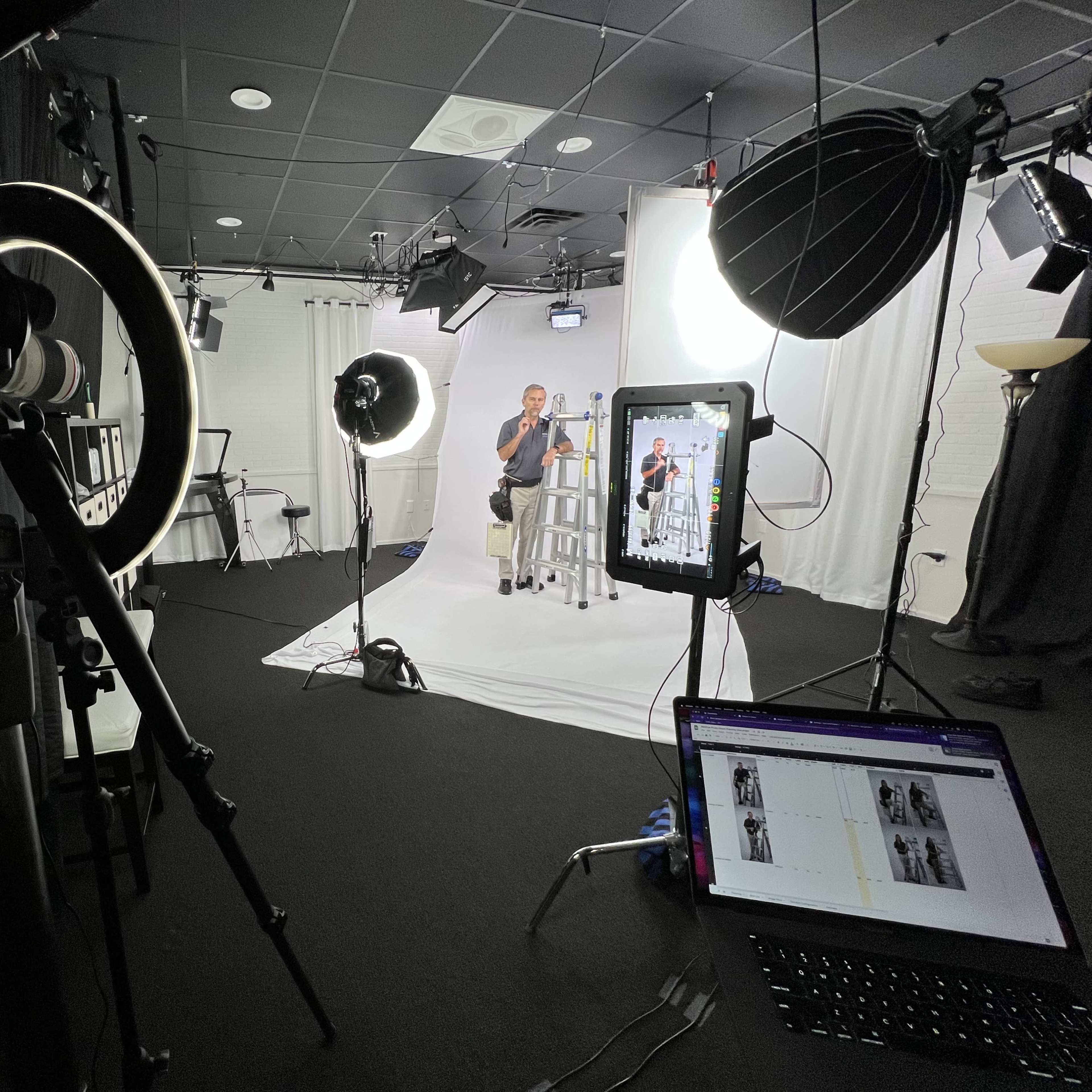 A photo studio setup features a seated subject under bright lights, with camera equipment, a laptop displaying images, and various lighting tools arranged around a white backdrop.