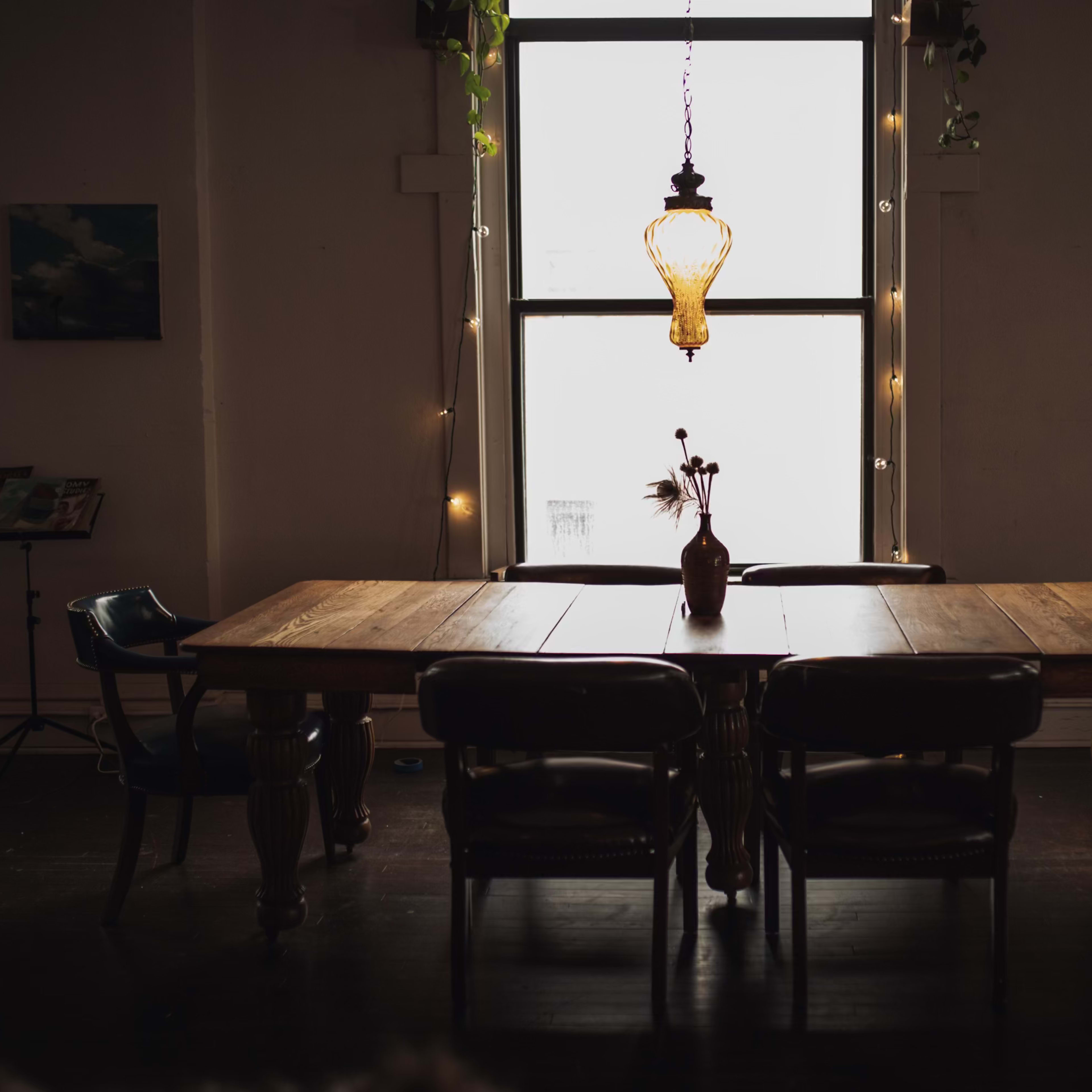 A wooden dining table surrounded by chairs is illuminated by a hanging light fixture in a dimly lit room.