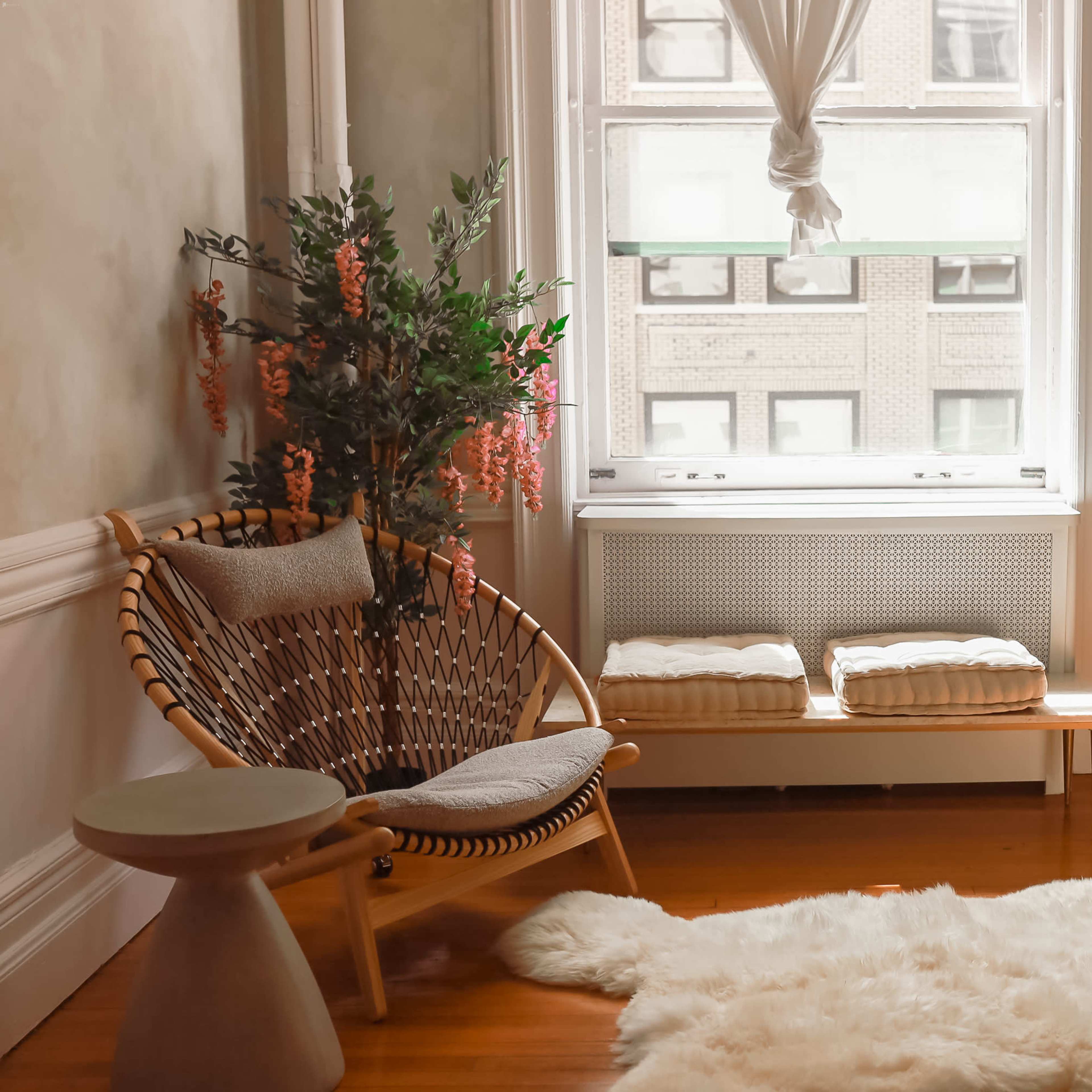 A cozy corner features a rattan chair, a small table, a bench, and lush greenery by a window.