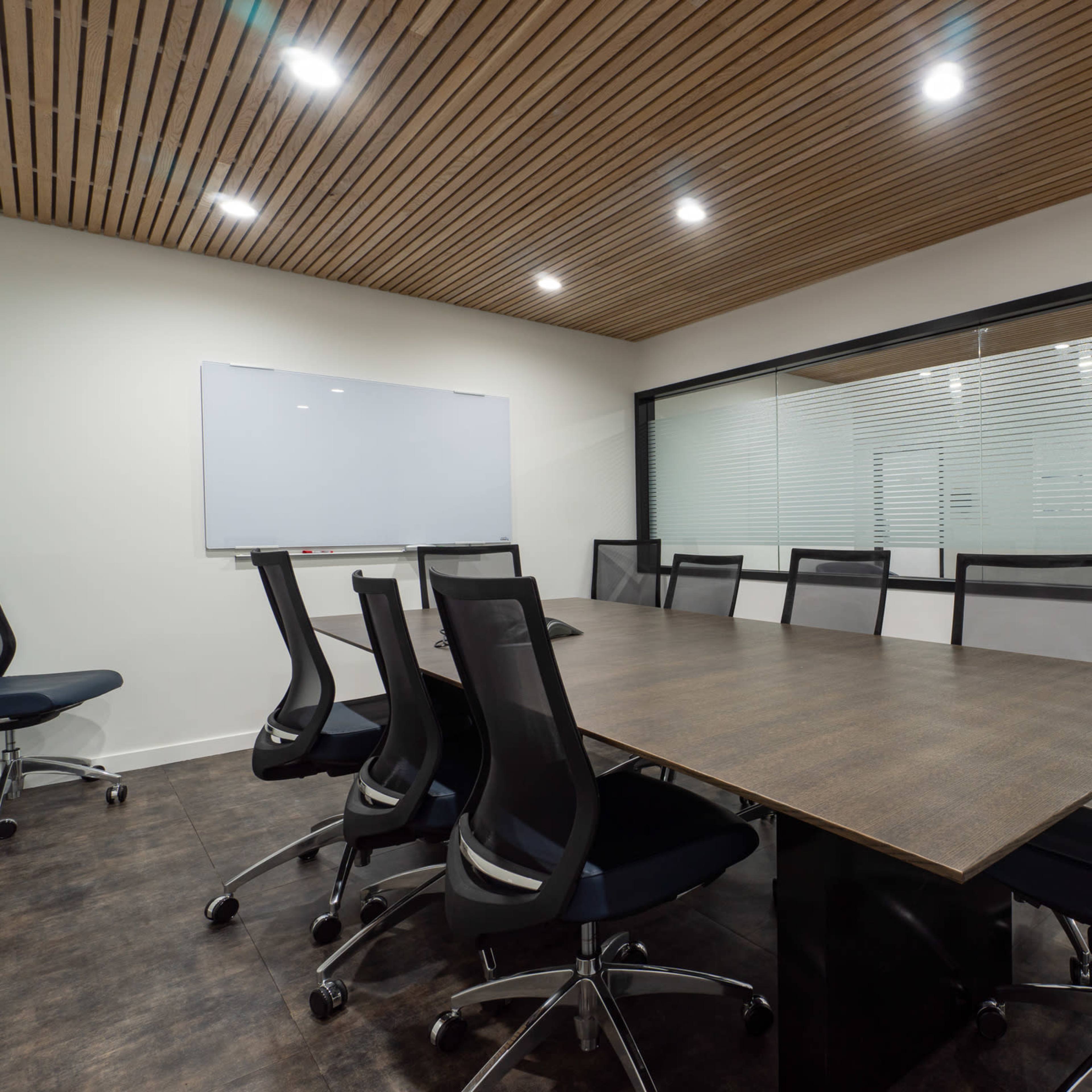 A conference room features a long wooden table surrounded by black ergonomic chairs, with a whiteboard and glass partition in the background.