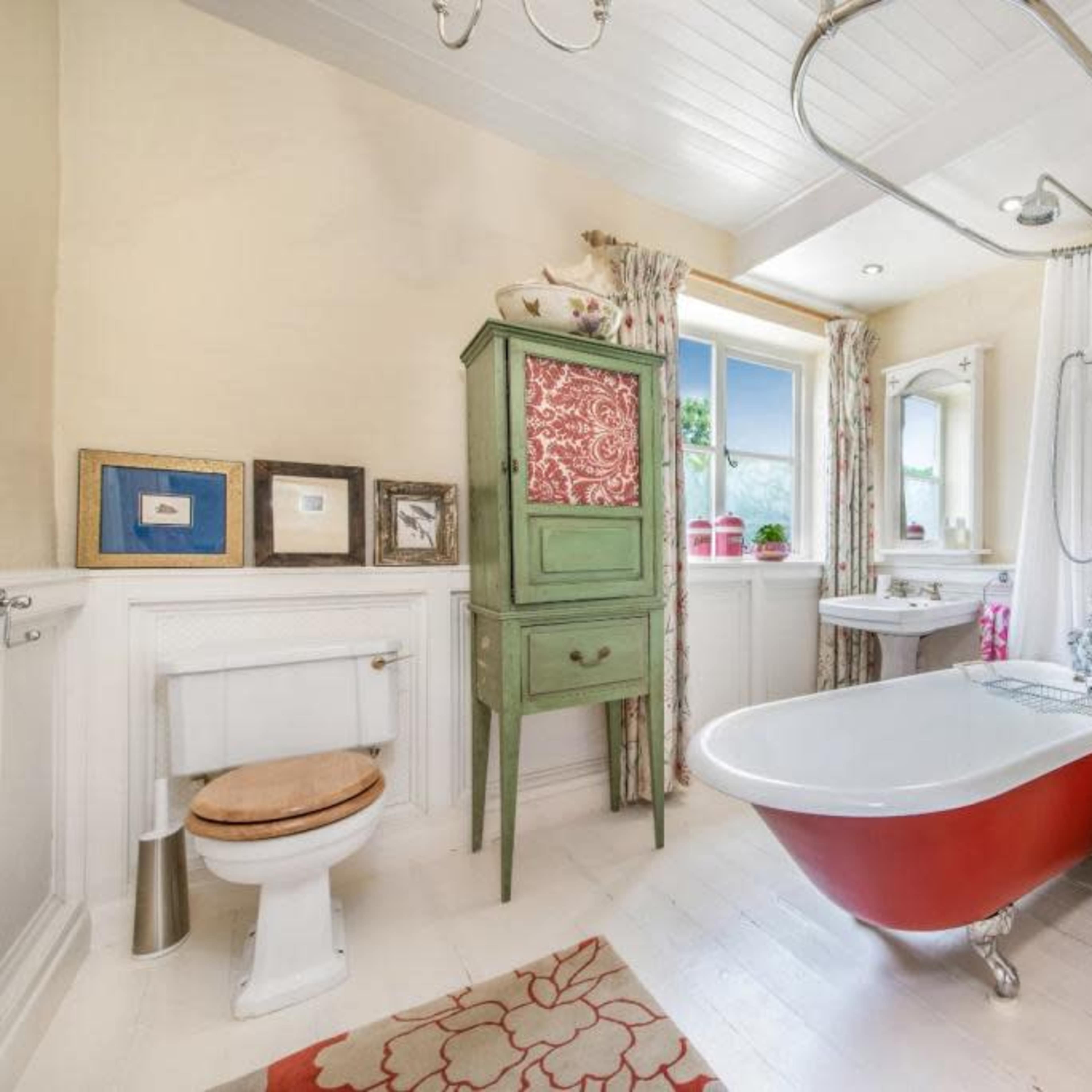 A bathroom featuring a freestanding red and white bathtub, a green cabinet, a window with curtains, and a white sink.