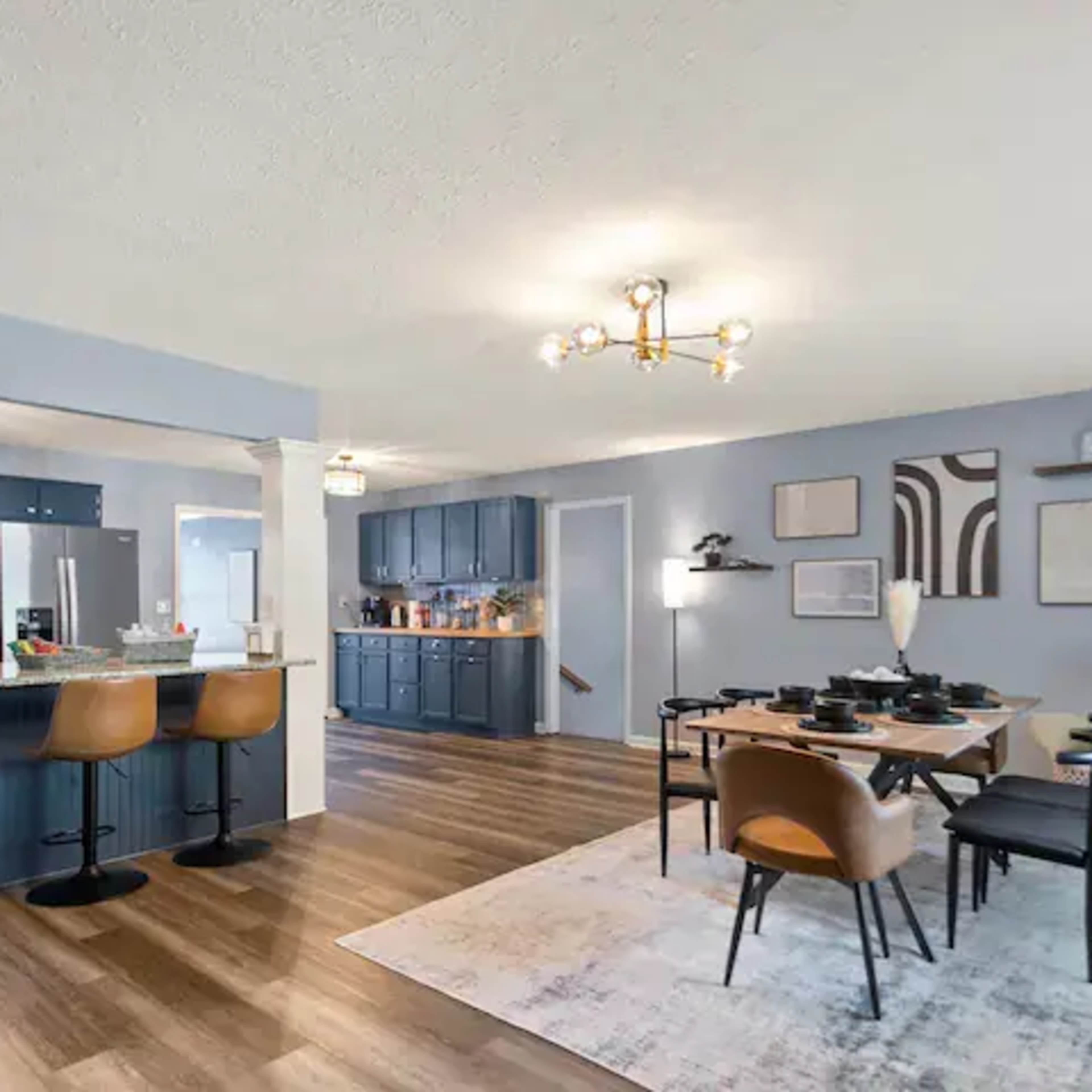 A modern kitchen and dining area feature blue cabinetry, a large island with bar stools, and a dining table set with dishware, all accented by wooden flooring and contemporary decor.