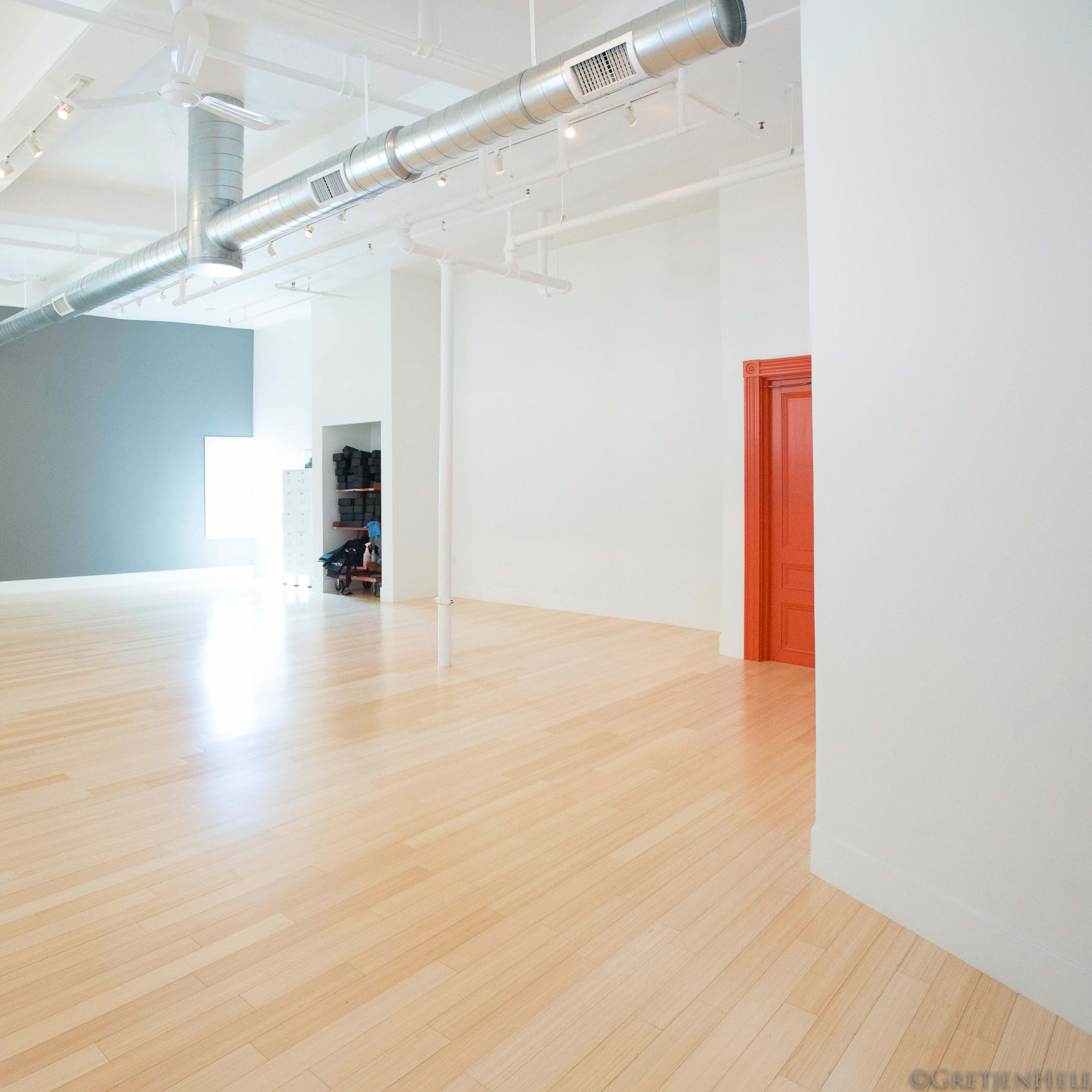 A spacious, empty room with light wood flooring, a gray accent wall, and a bright orange door.