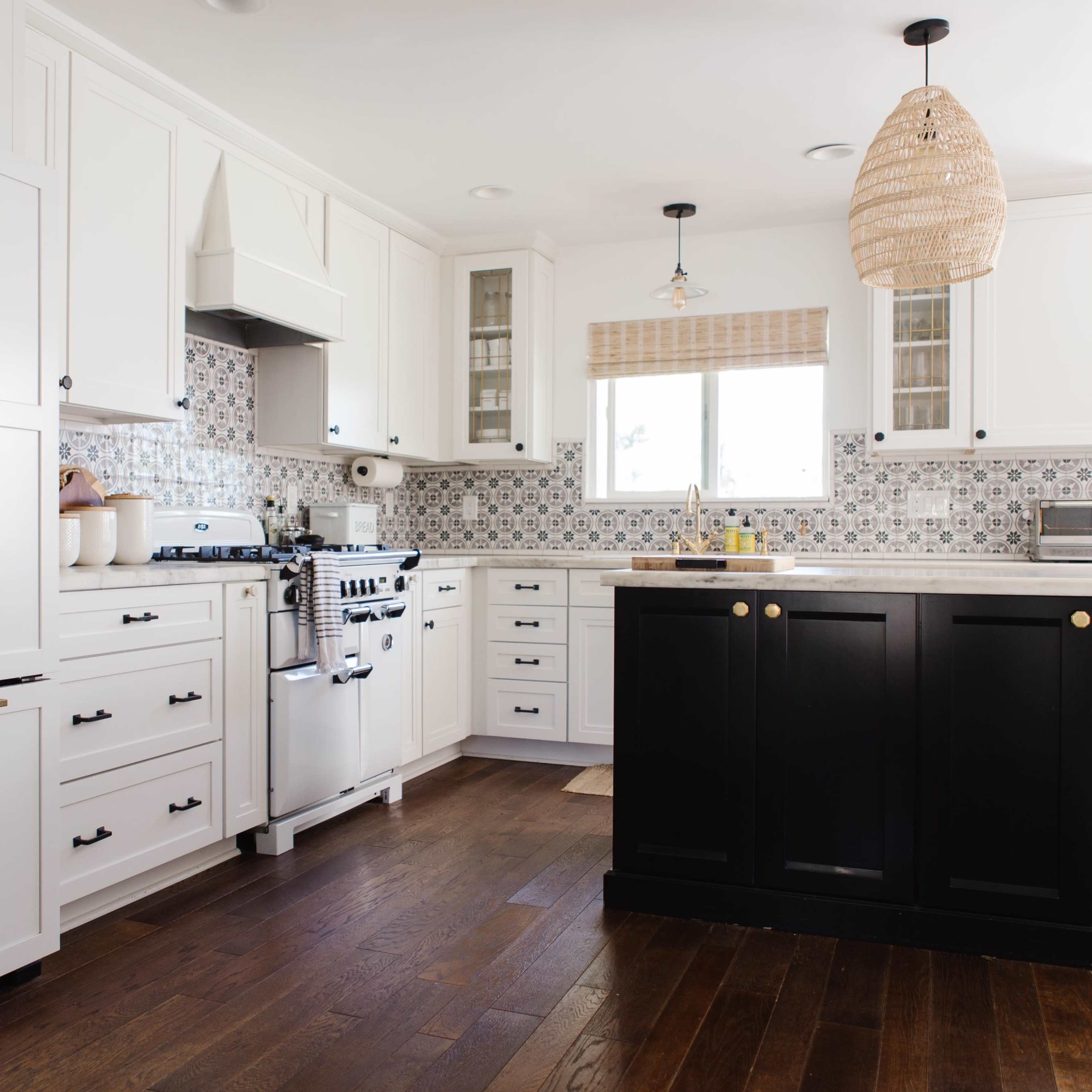 A modern kitchen featuring white cabinets on one side and black cabinets on the island, with a patterned backsplash and hardwood flooring.