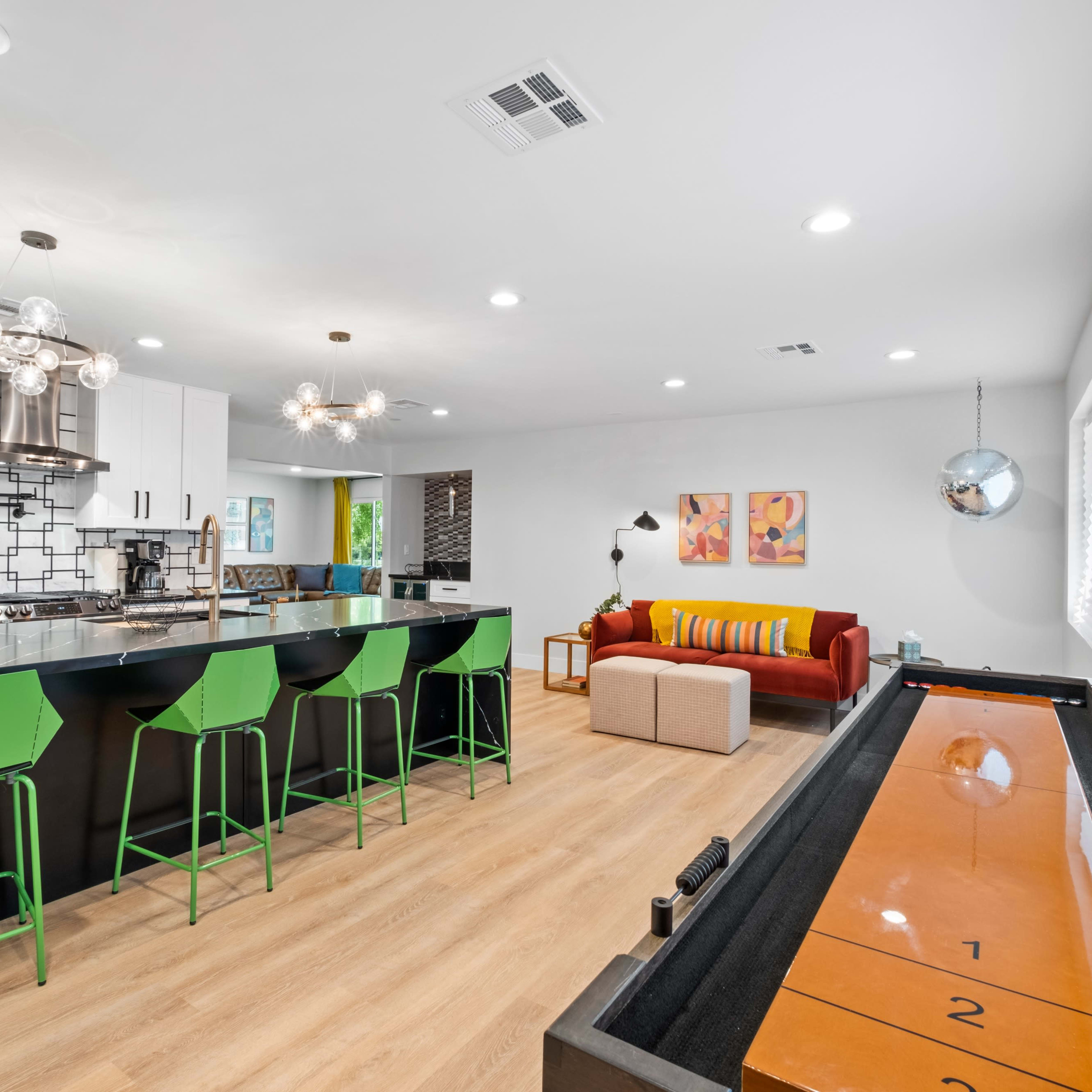 A modern kitchen with a large island, bright green bar stools, and a shuffleboard table in an open living space featuring a vibrant sofa and colorful artwork on the walls.