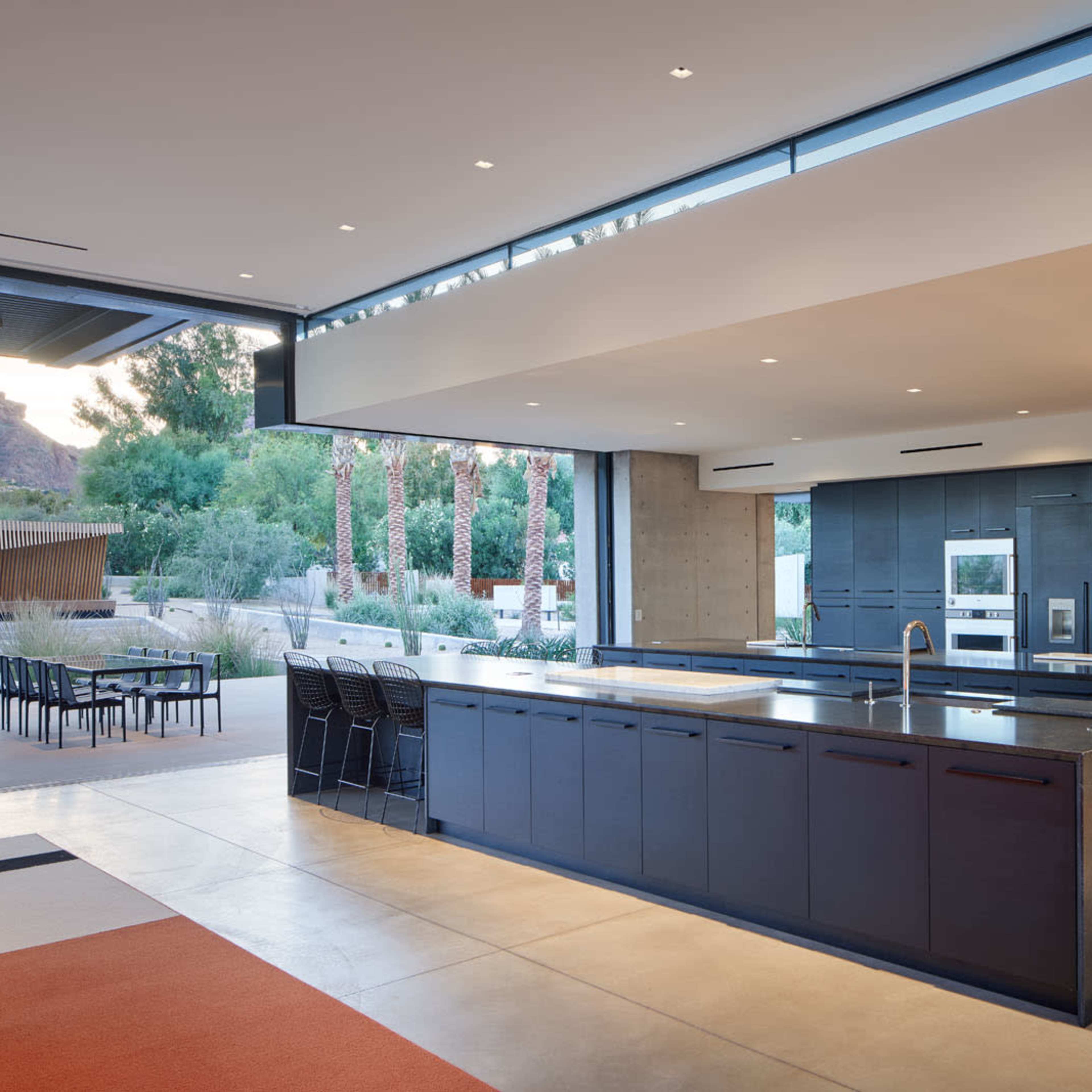 A modern kitchen with a large black island, concrete flooring, and floor-to-ceiling windows overlooking a patio and palm trees.