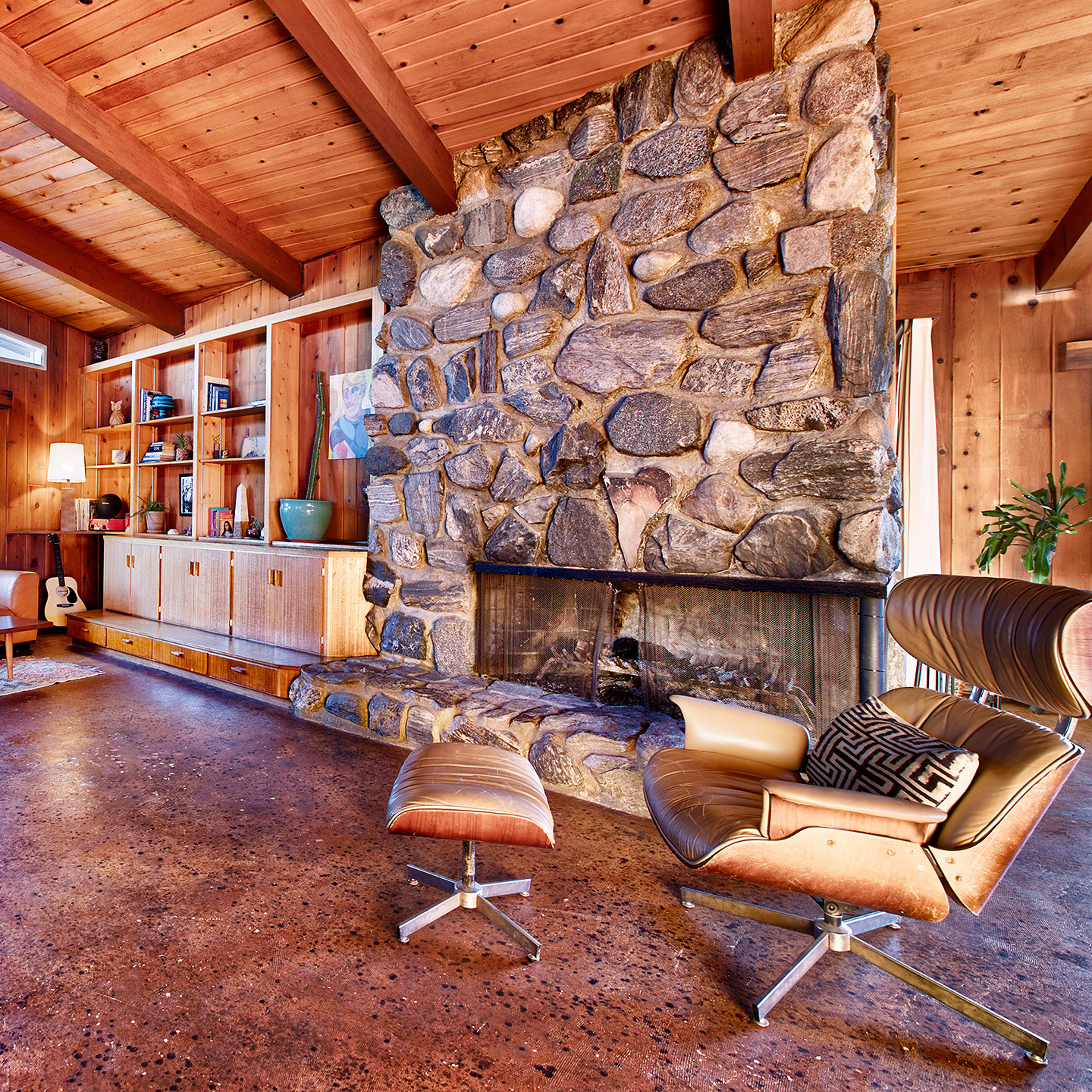 A spacious living room features a stone fireplace, wooden beams, and modern furniture arranged around a central coffee table.