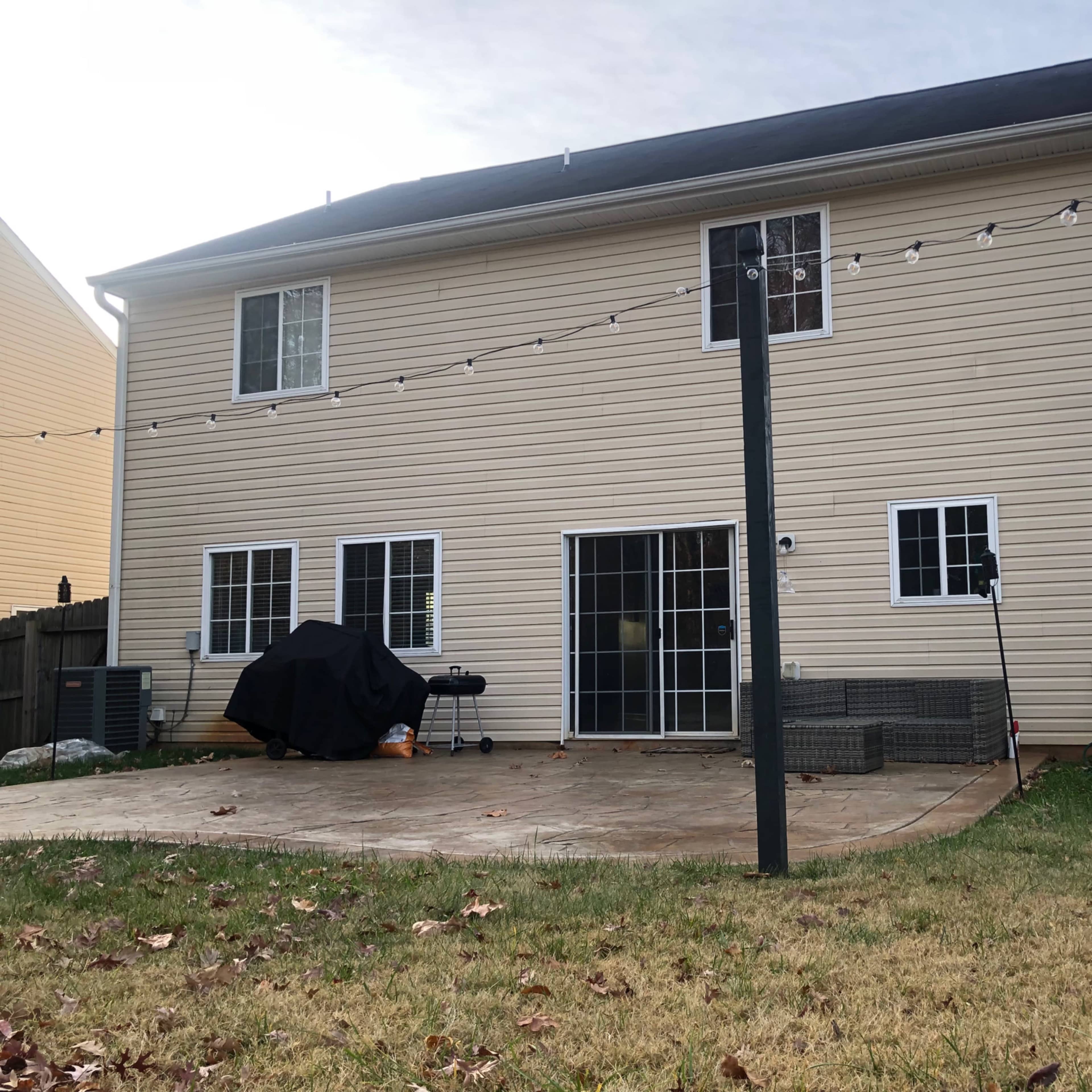 The back of a two-story house with a patio area and outdoor furniture, along with a grill and string lights overhead.