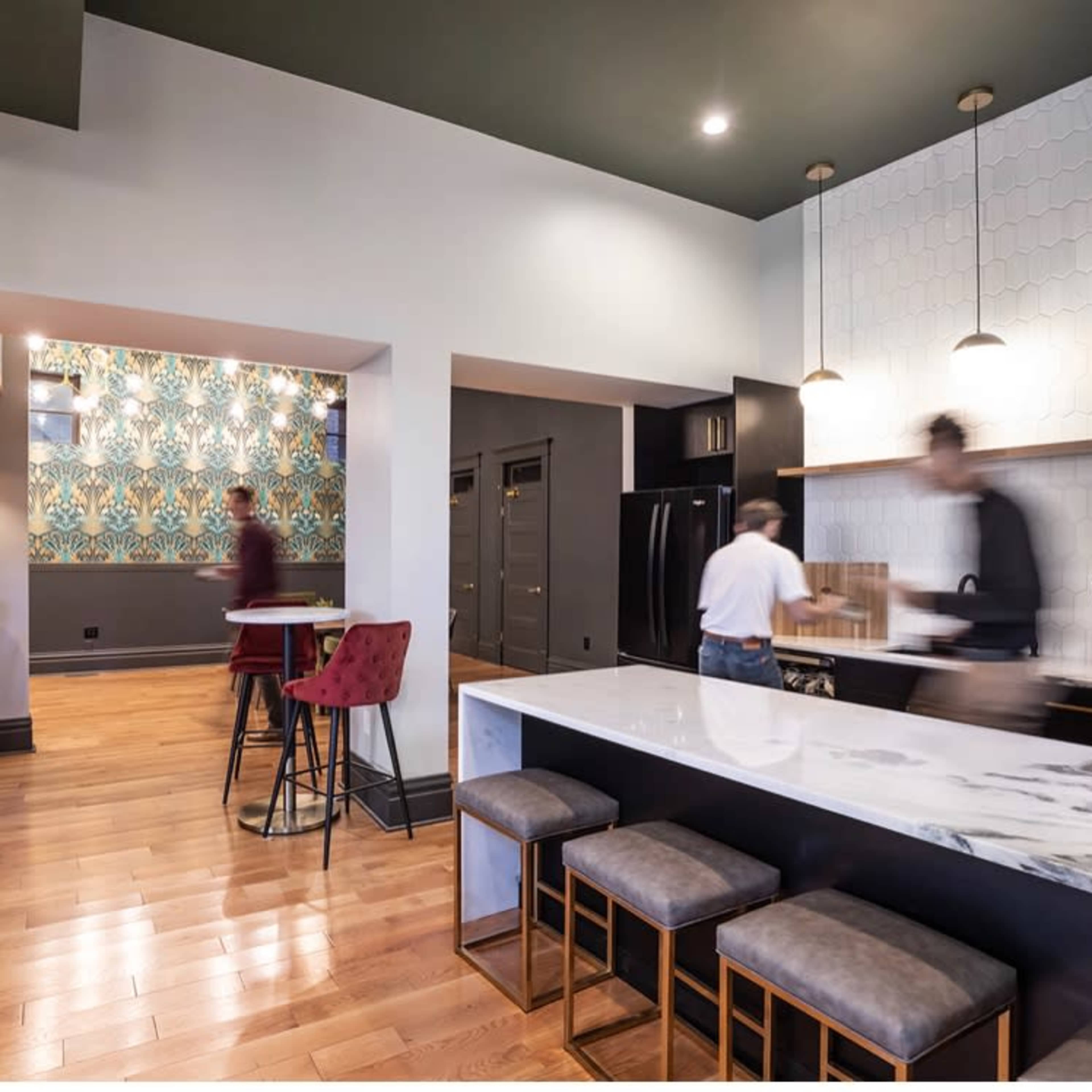 A modern kitchen with a large marble island, wooden flooring, and a patterned accent wall, while several people are engaged in activities in the space.