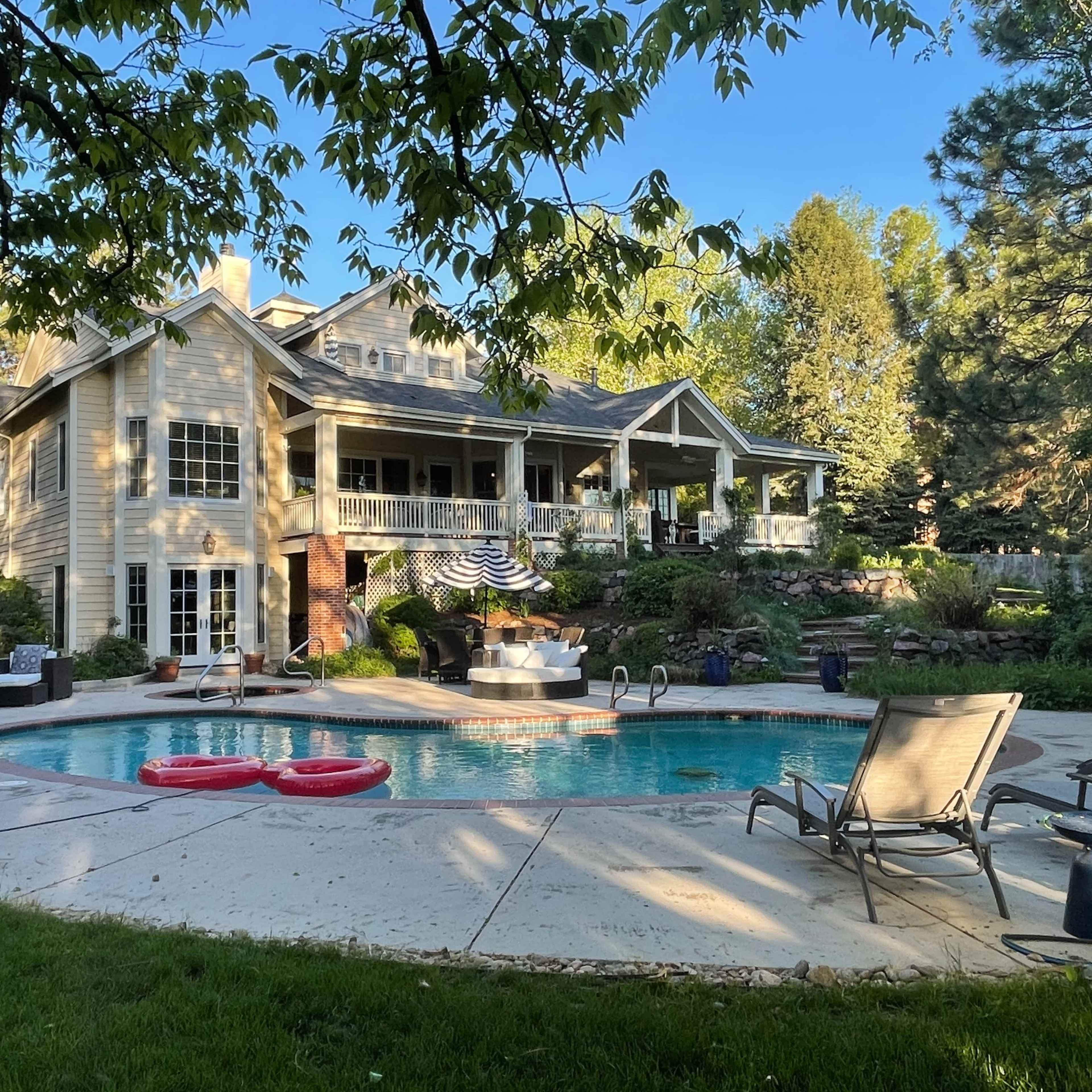 A spacious backyard features a swimming pool with lounge chairs and two inner tubes, surrounded by a two-story house and lush greenery.