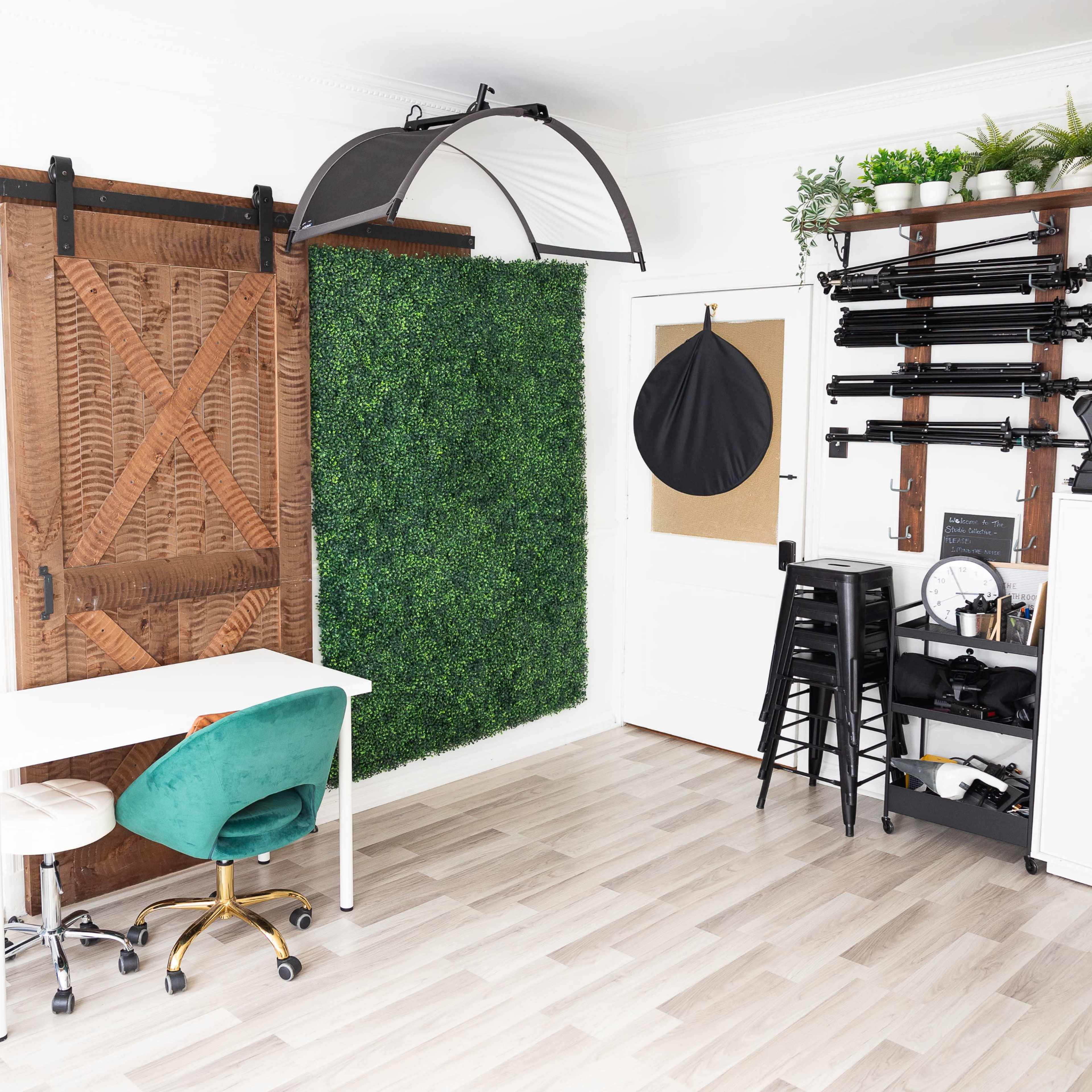 A modern workspace featuring a white desk with a green chair, a large wooden sliding door, a wall covered in artificial greenery, and various photography equipment mounted on the opposite wall.