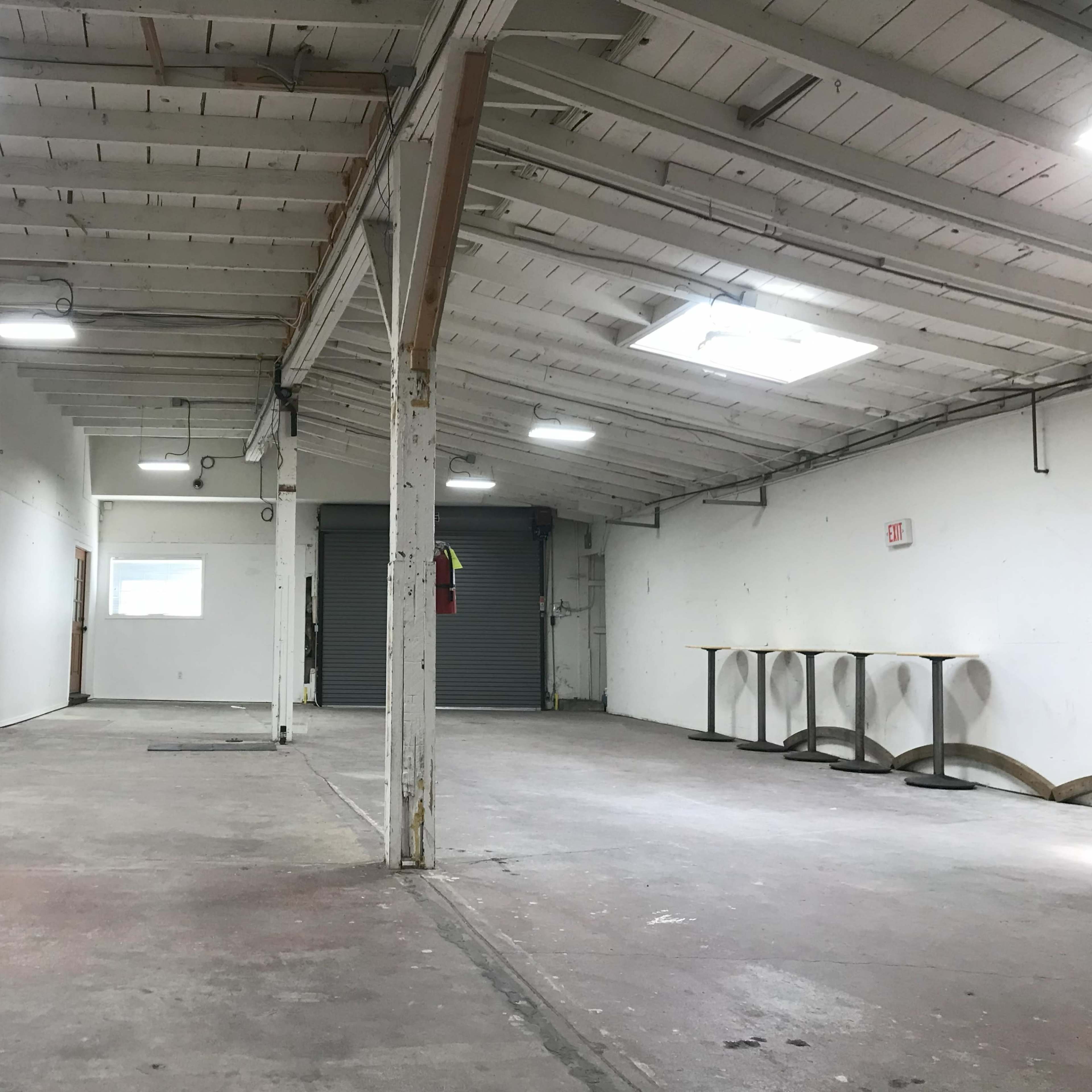 A vacant industrial space with high ceilings, exposed beams, and minimal furnishings, including a kitchenette along one wall.