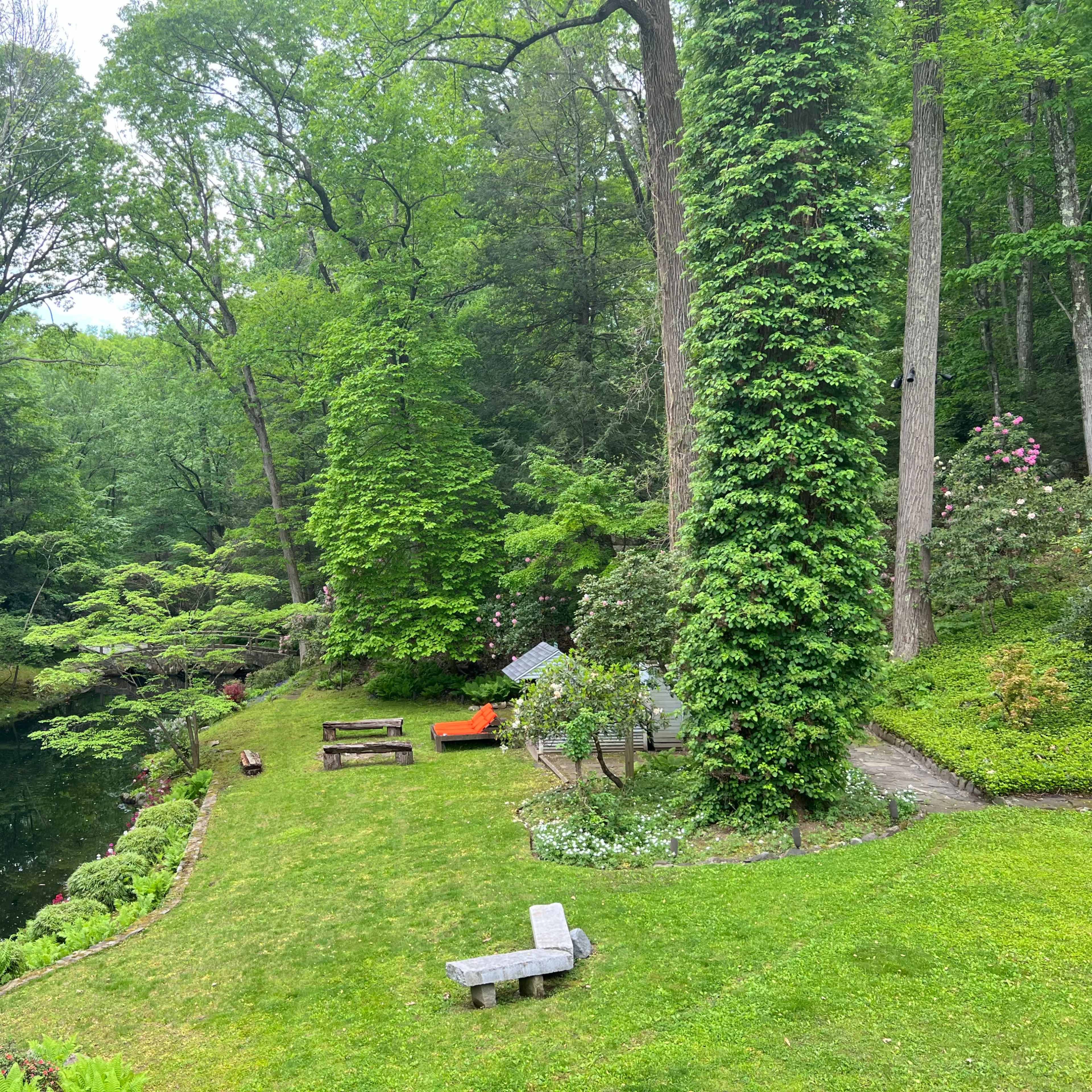 A lush green landscape with a pond, surrounded by trees, and a seating area with benches and an orange chair.