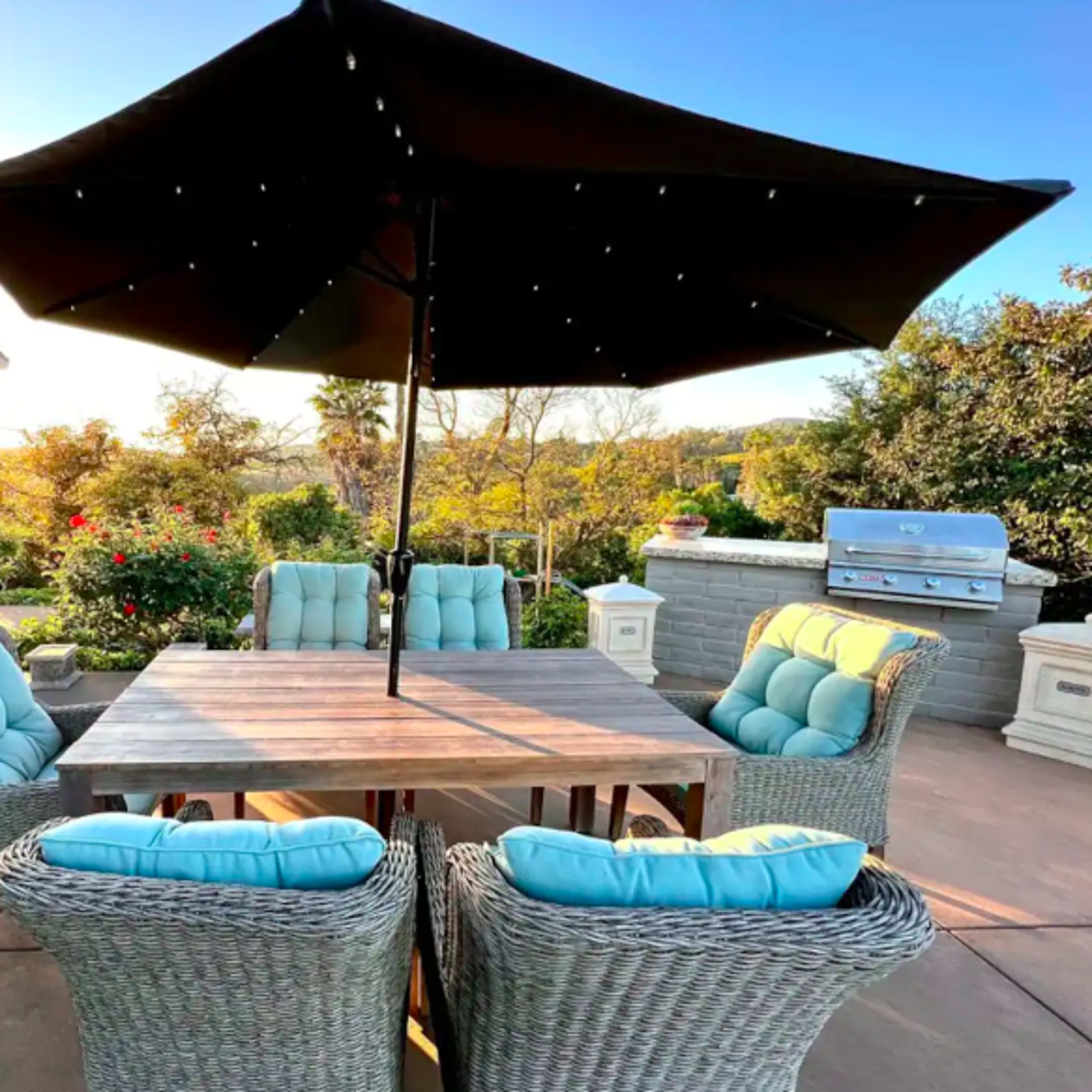 A patio features a wooden table surrounded by six wicker chairs with light blue cushions, shaded by a large umbrella, overlooking landscaped greenery and a barbecue grill.