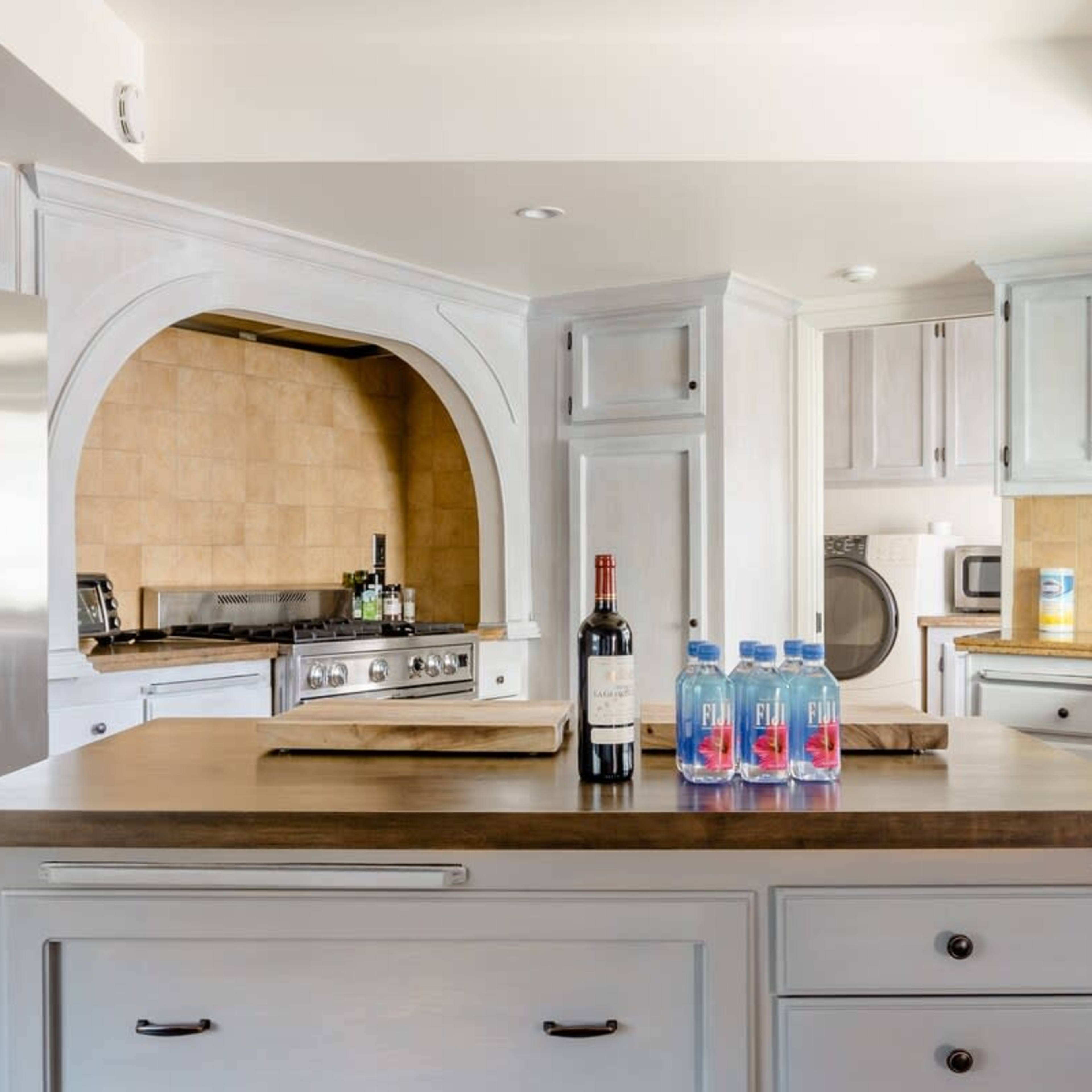 A spacious kitchen features a central island with wine and bottles of water, stainless steel appliances, and light-colored cabinetry.