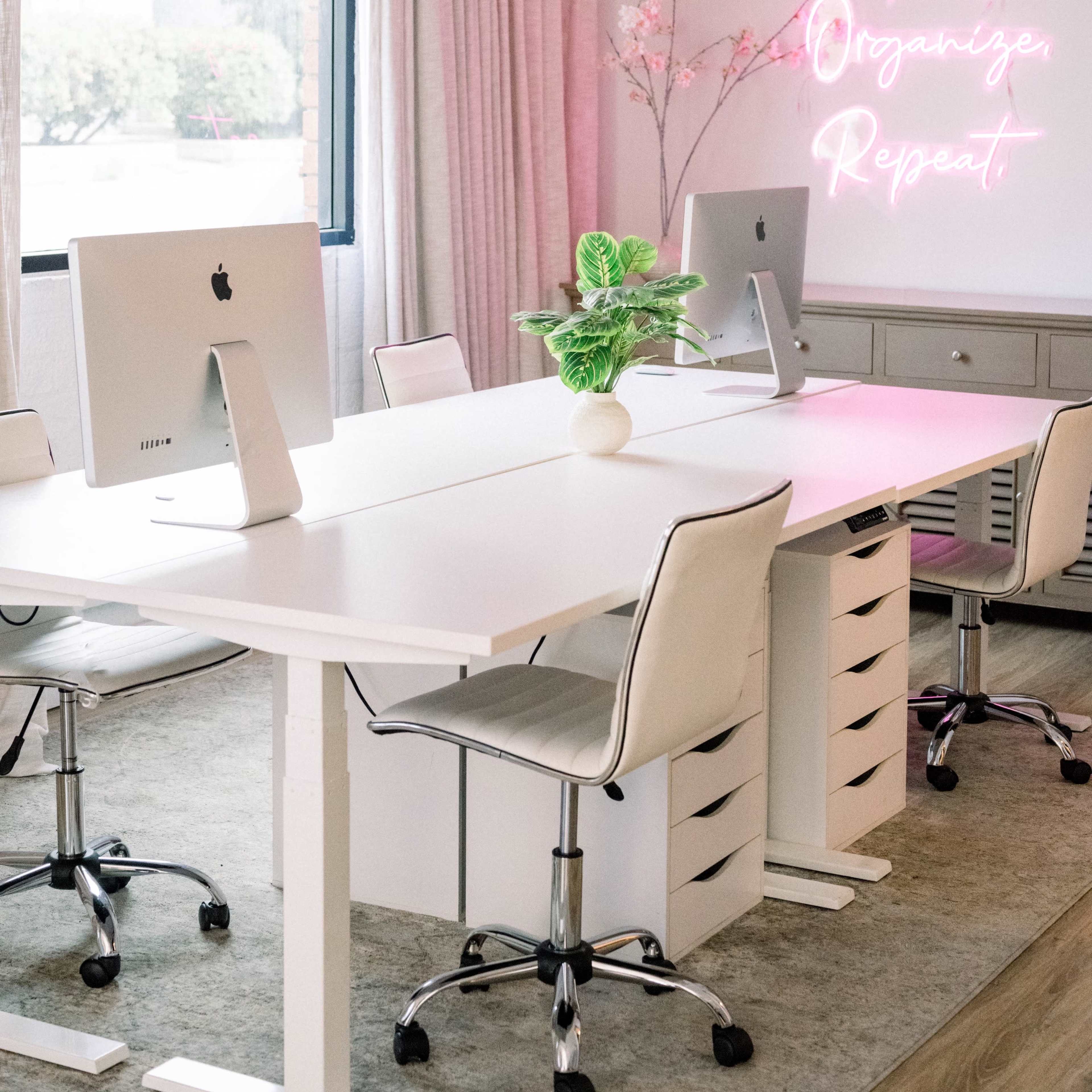 A modern office space with two computers on white desks, a plant centerpiece, and a neon sign that reads "Play Organizer Repeat."