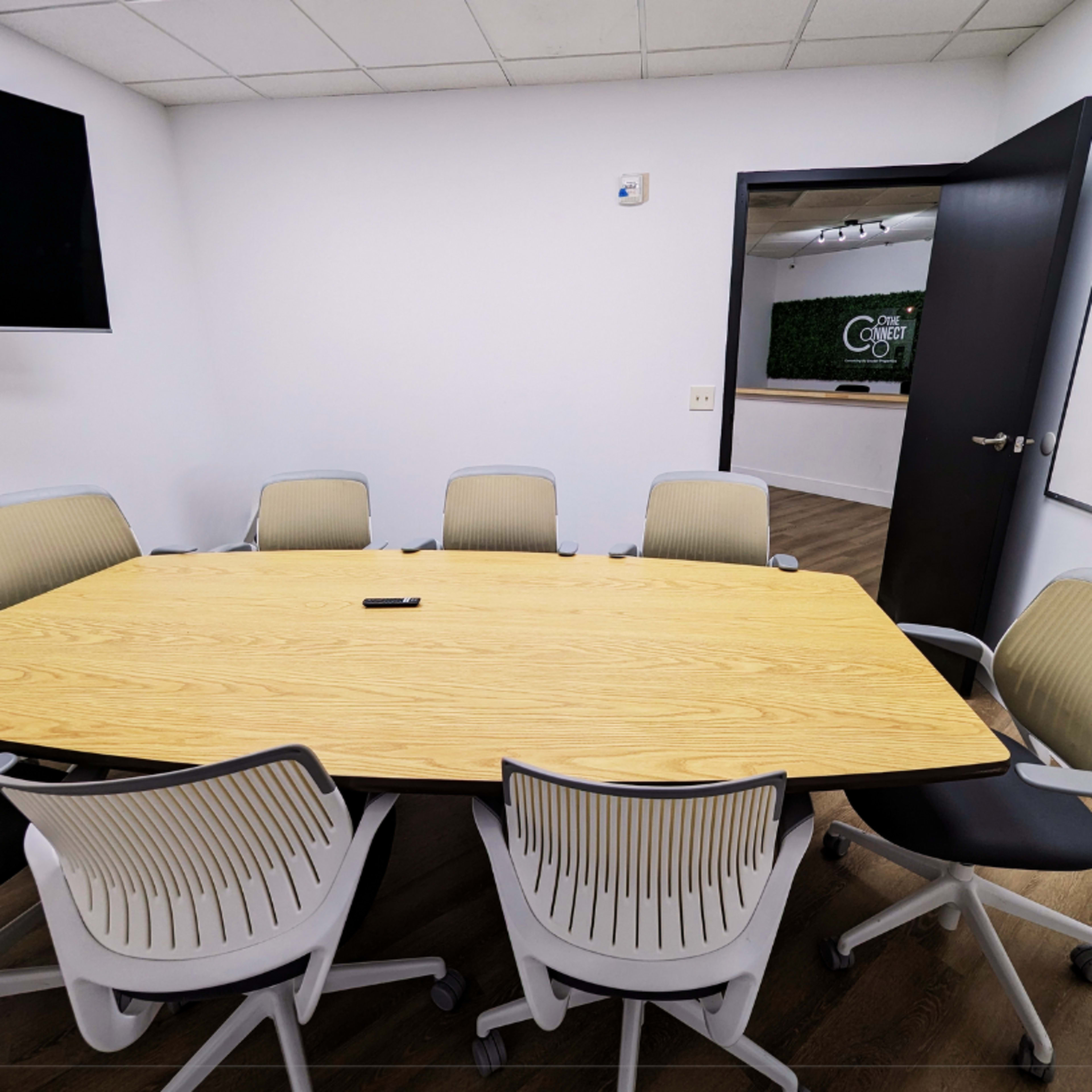 A large, oval wooden conference table surrounded by white and gray office chairs is set in a minimalist meeting room.