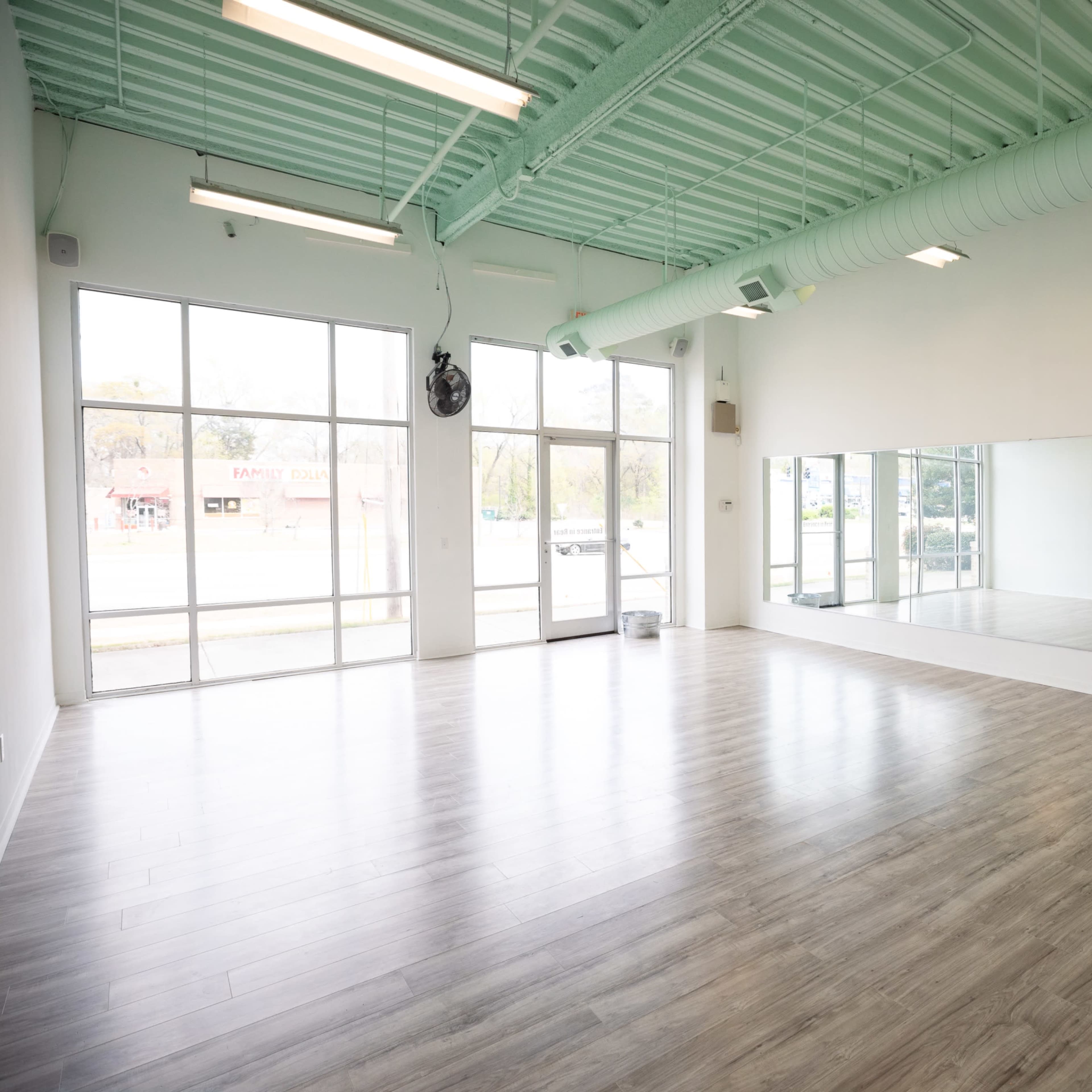 A spacious, empty studio with large windows, a mirror along one wall, and light wooden flooring.