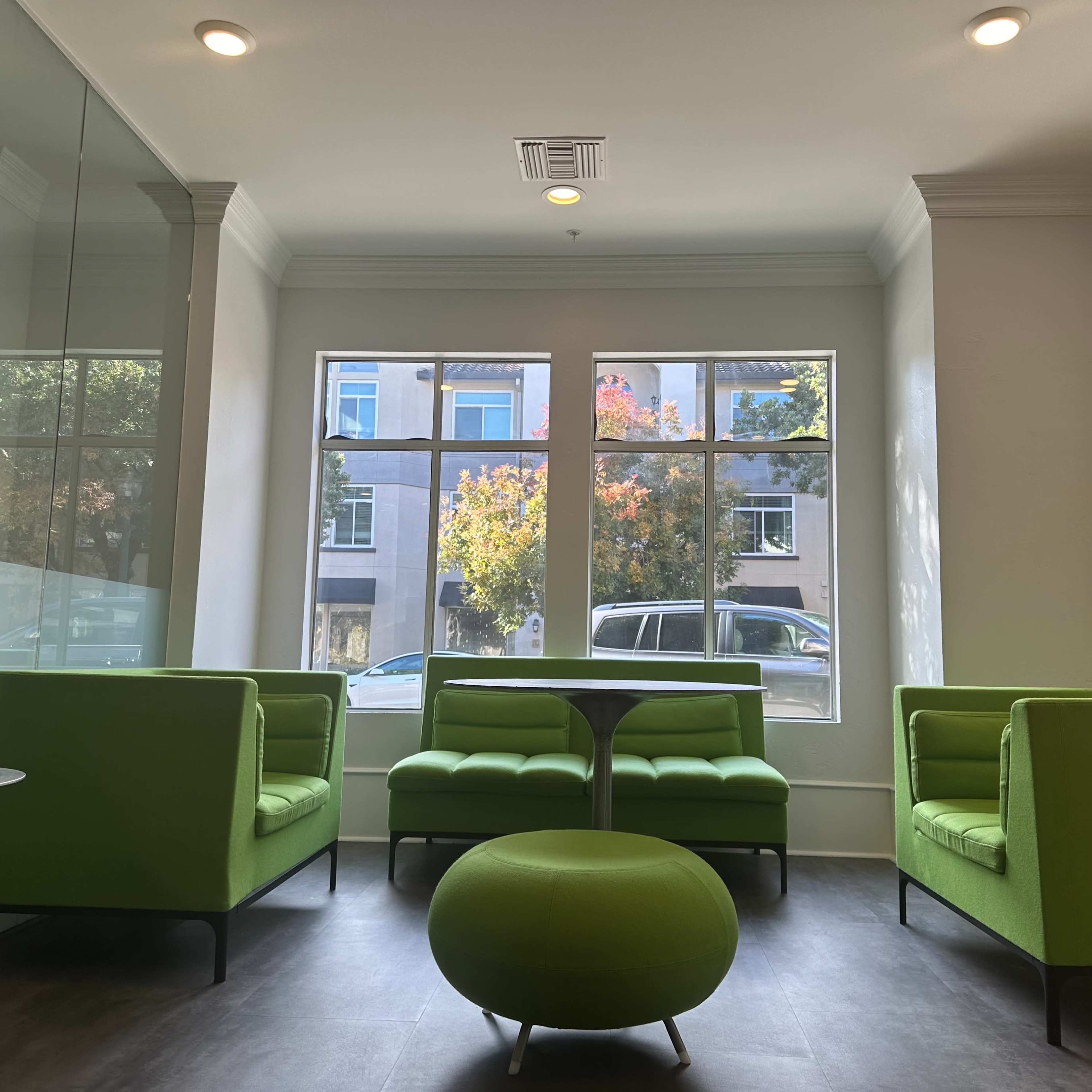 A modern waiting area with green seating, large windows allowing natural light, and a round green table.