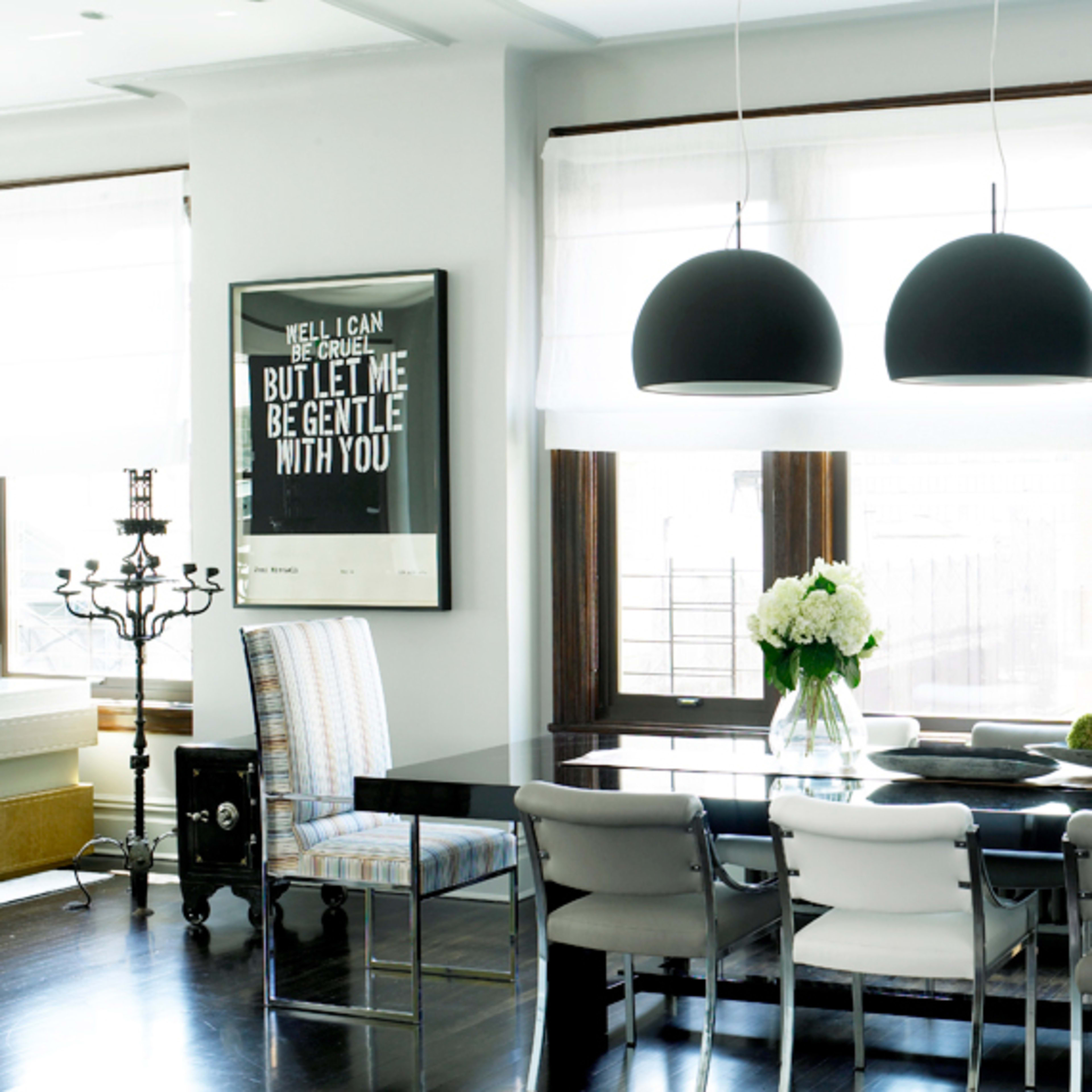 A modern dining room features a long black table surrounded by various chairs, with three pendant lights hanging above and a decorative floral arrangement on the table.