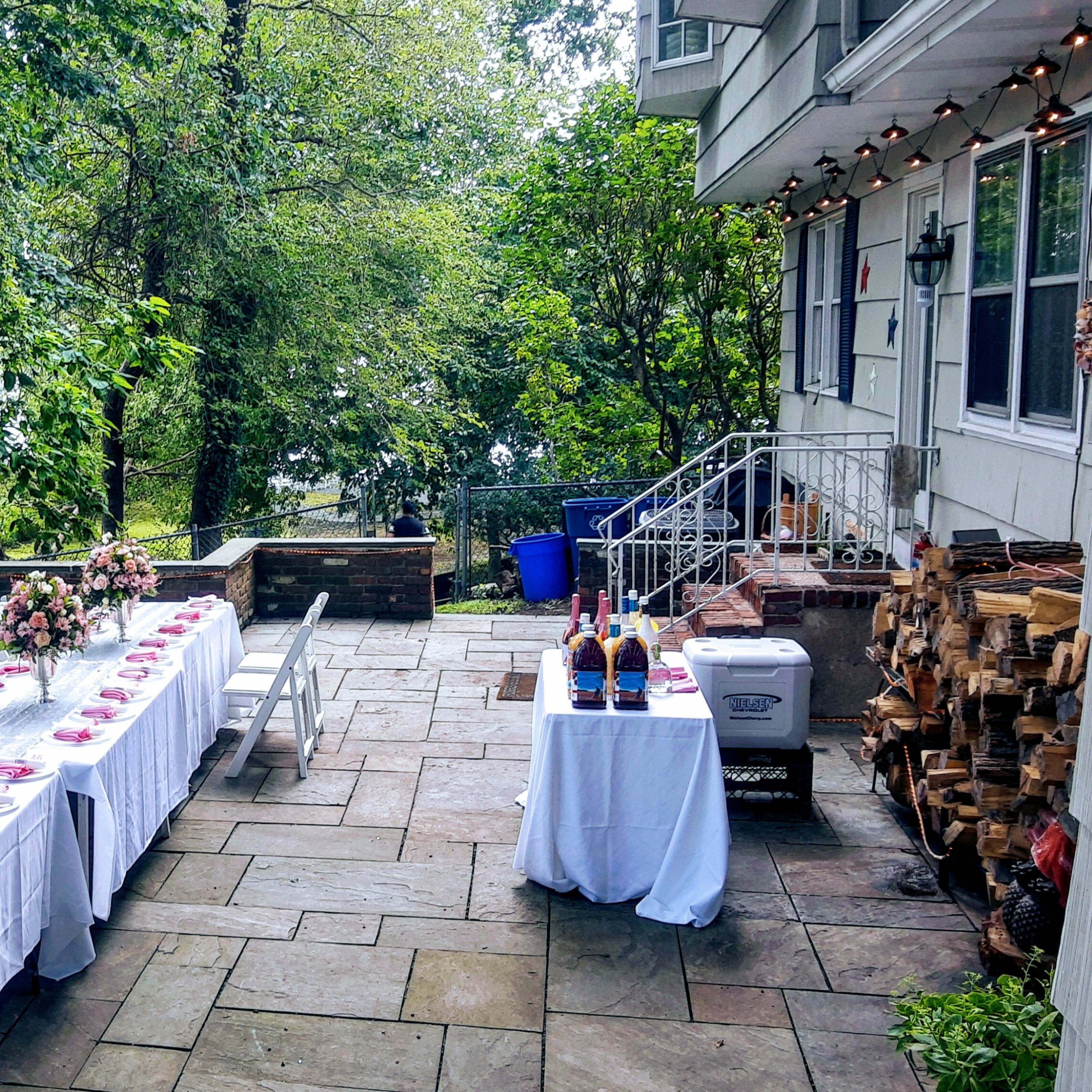 A long table set with pink plates and floral arrangements is positioned on a stone patio surrounded by greenery and a view of the water.