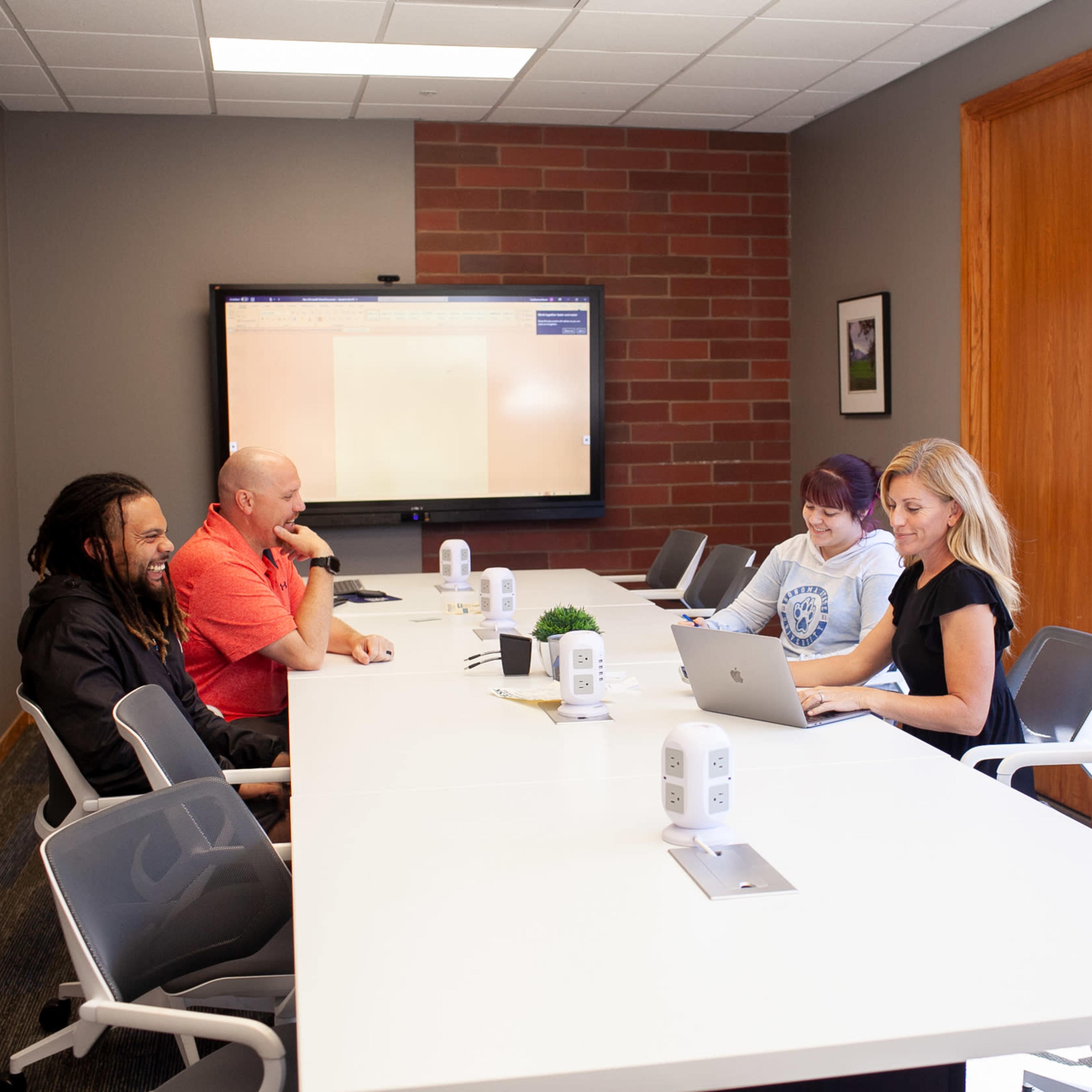 A group of four people sit around a long table in a conference room, with a screen displaying content in the background.