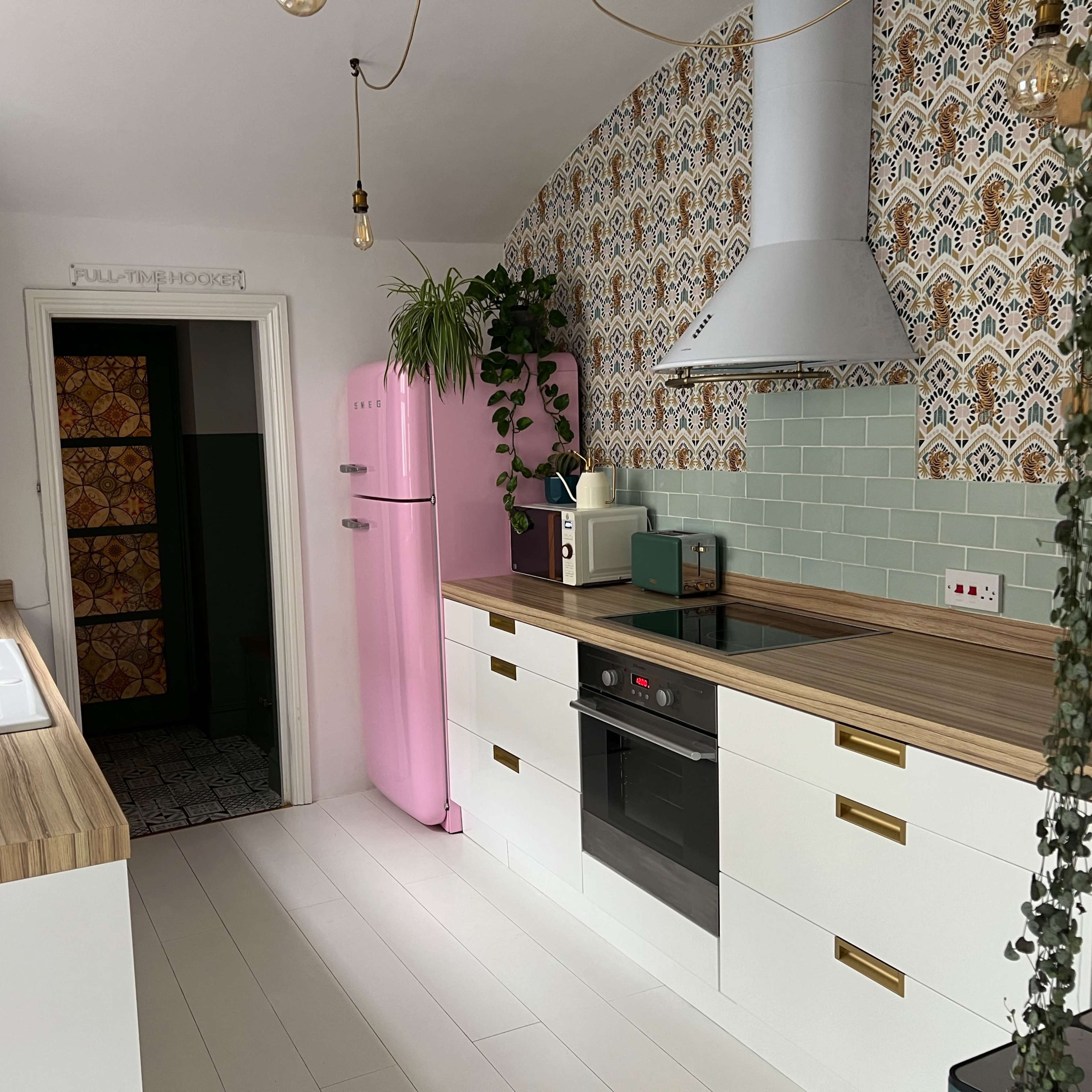 A modern kitchen features a pink refrigerator, white cabinetry, wooden countertops, and patterned wallpaper.