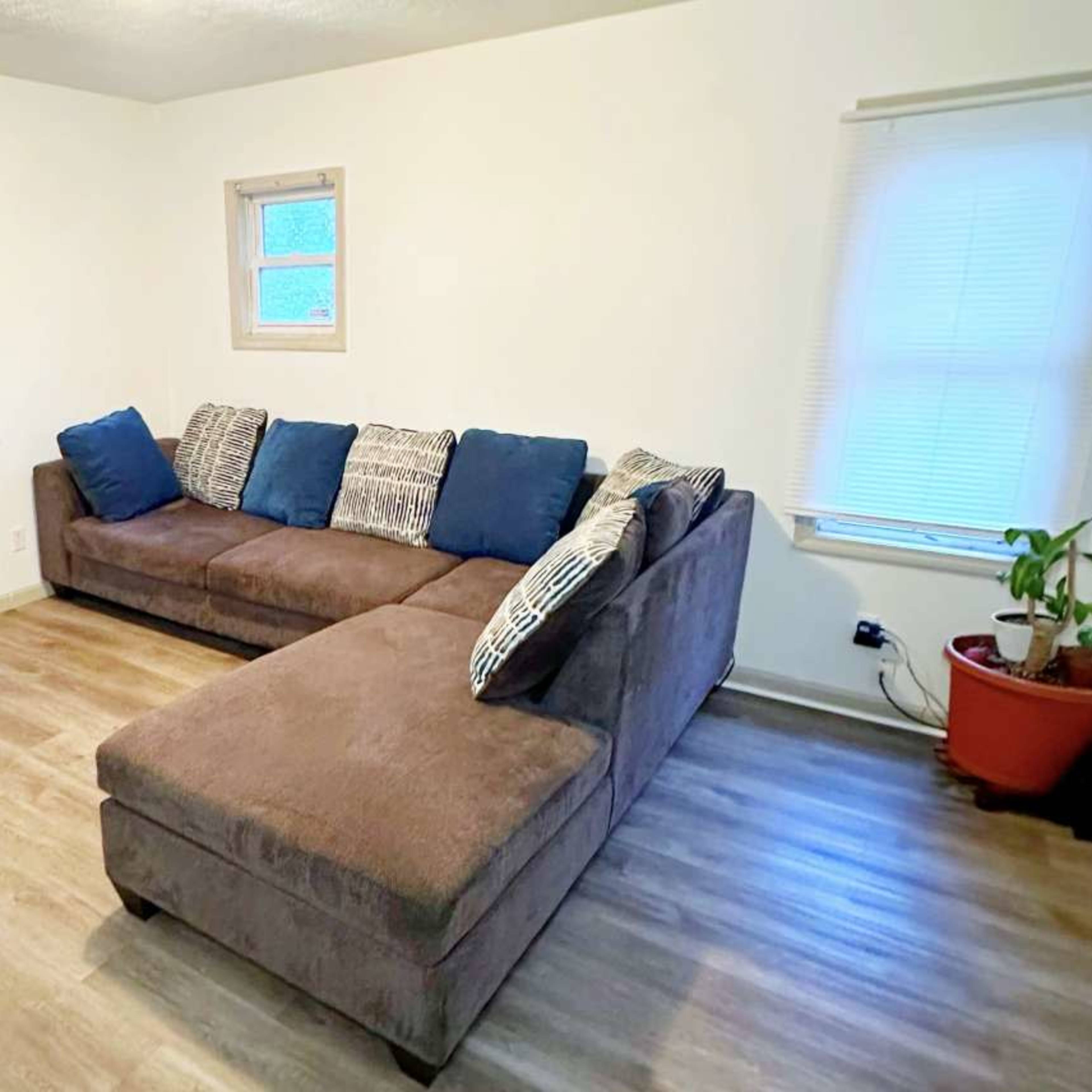 A brown sectional sofa with blue and patterned pillows sits in a bright room featuring a window and a potted plant.