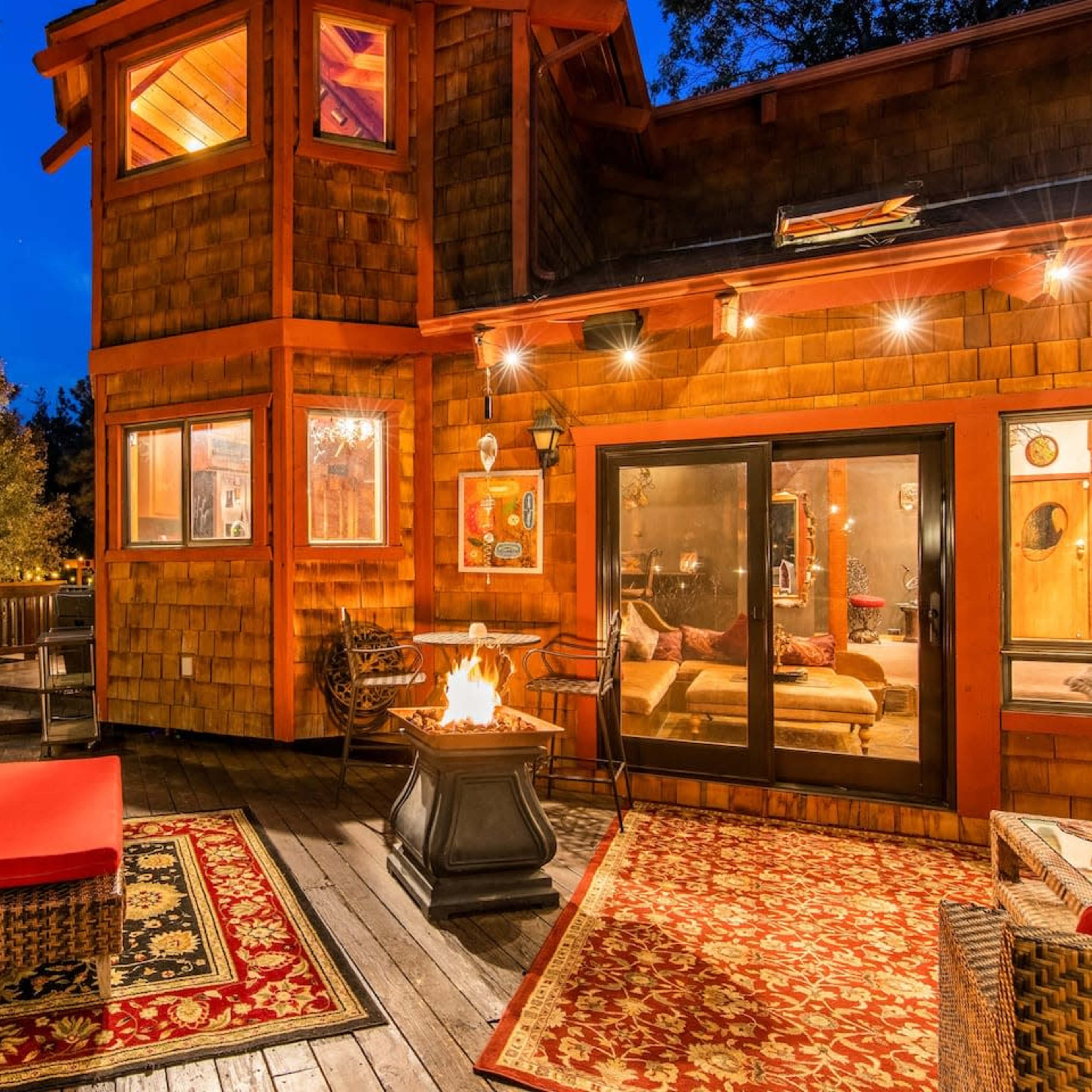 A wooden deck features a seating area with a fire pit and decorative rugs, surrounded by trees and illuminated by string lights against a twilight sky.