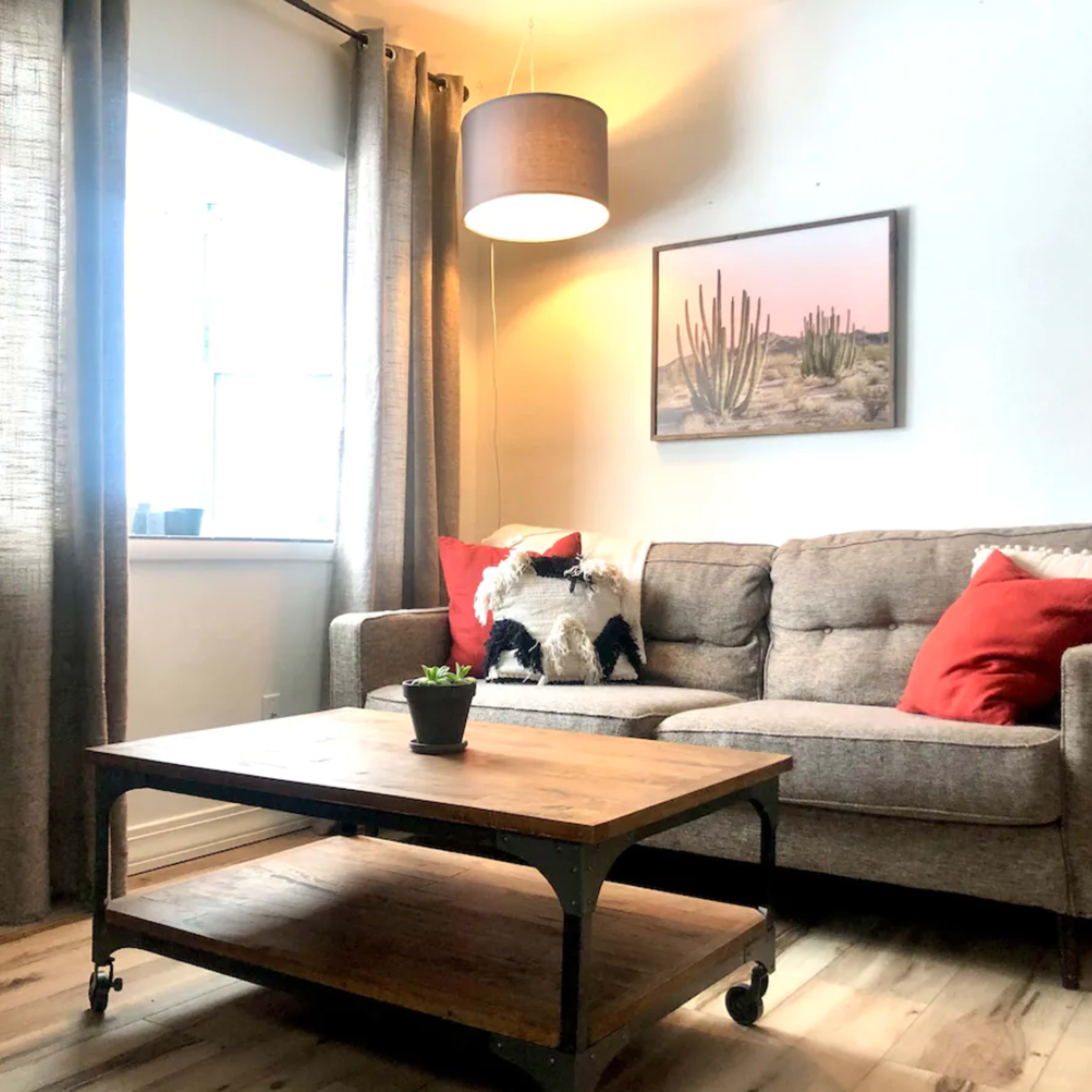 A living room features a gray sofa with red pillows, a wooden coffee table, and a large window adorned with curtains, illuminated by a hanging light.