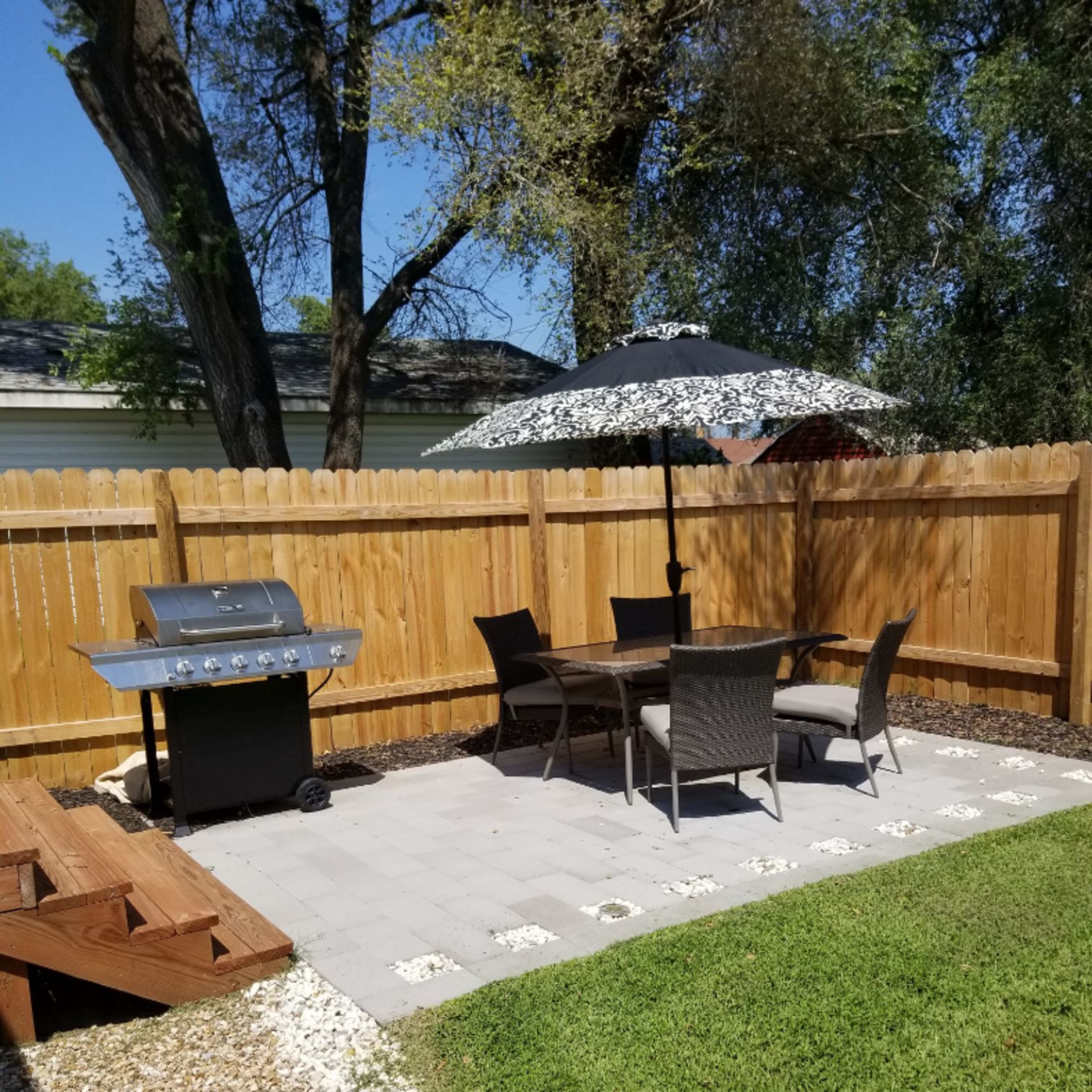 A backyard patio features a grill, dining table with chairs, and an umbrella, surrounded by a wooden fence and landscaped with stones and grass.