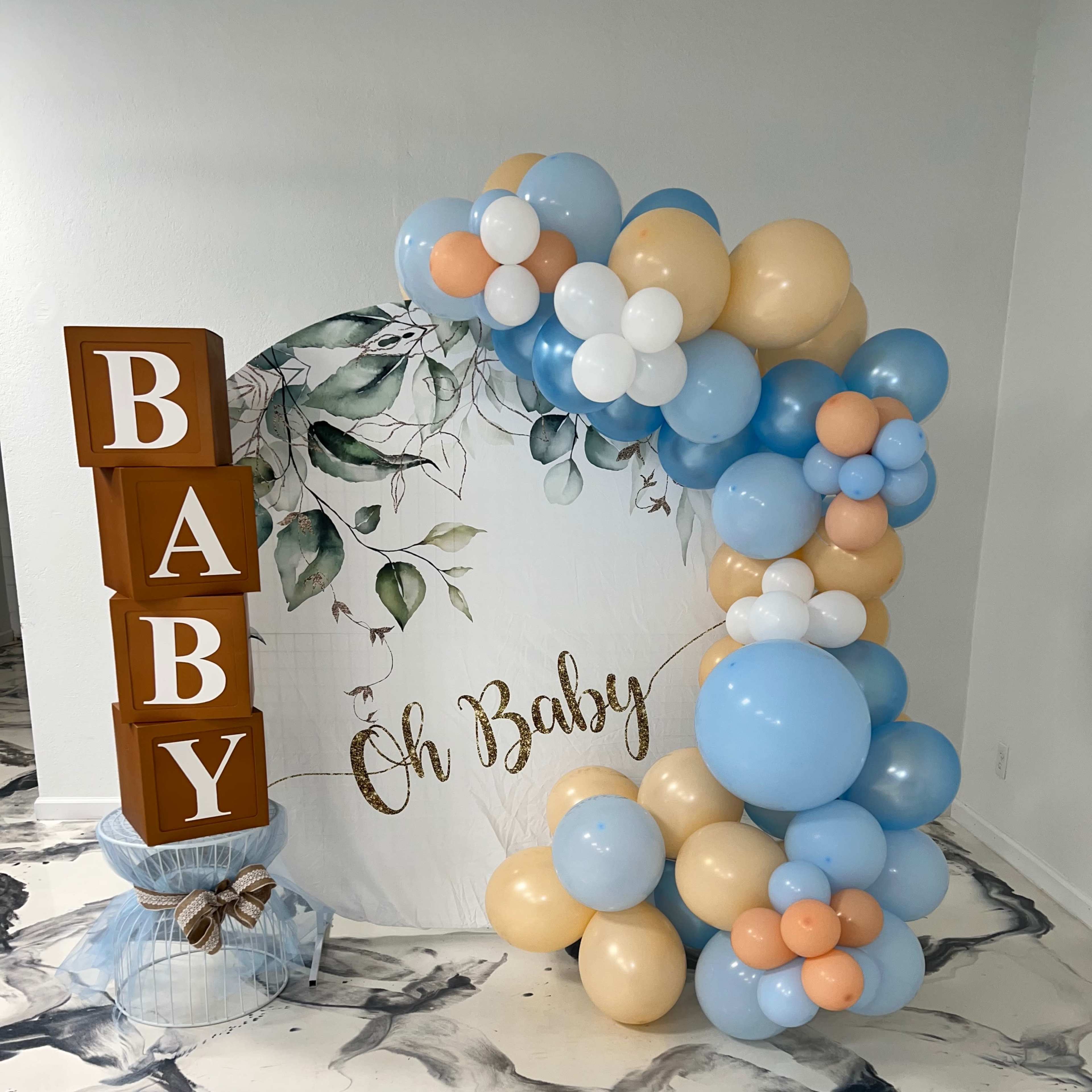 A decorative setup for a baby shower, including a large sign that reads "Oh Baby" and an arch made of blue, white, and beige balloons.