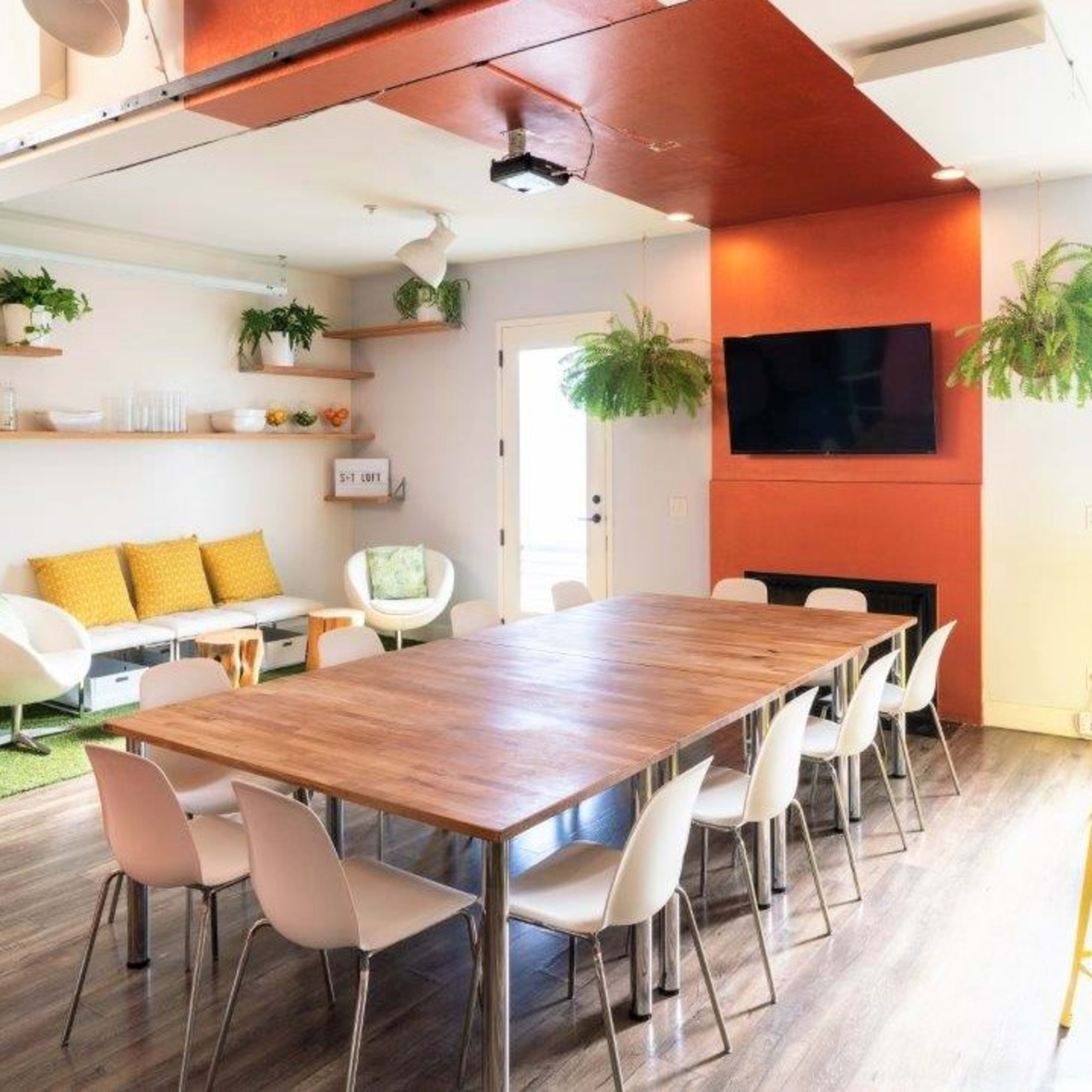 A modern communal space featuring a long wooden table surrounded by chairs, a cozy seating area with a sofa and plants, and bright yellow tables in a well-lit environment.