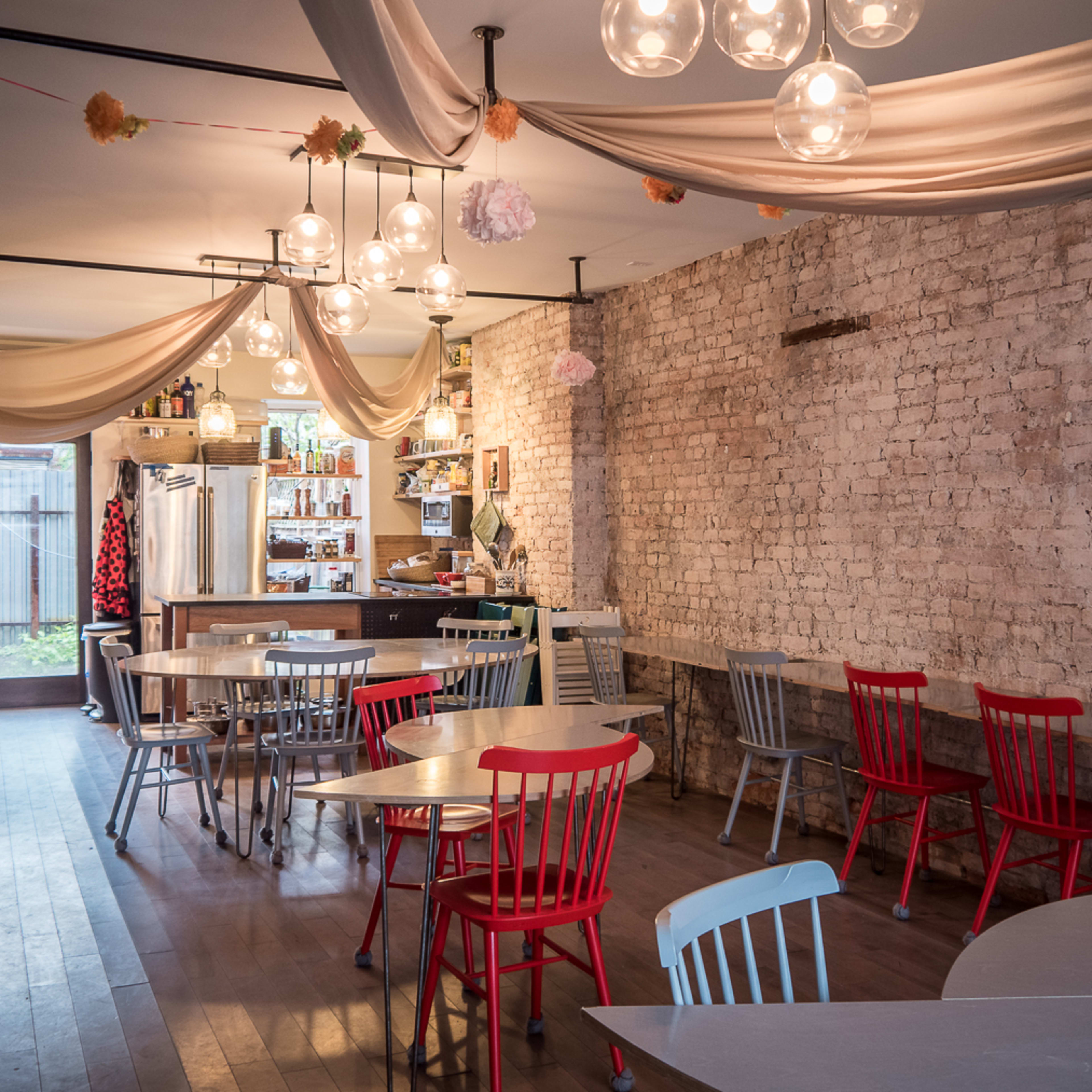A rustic café interior featuring a mix of red and gray chairs around circular tables, with exposed brick walls and decorative lighting.
