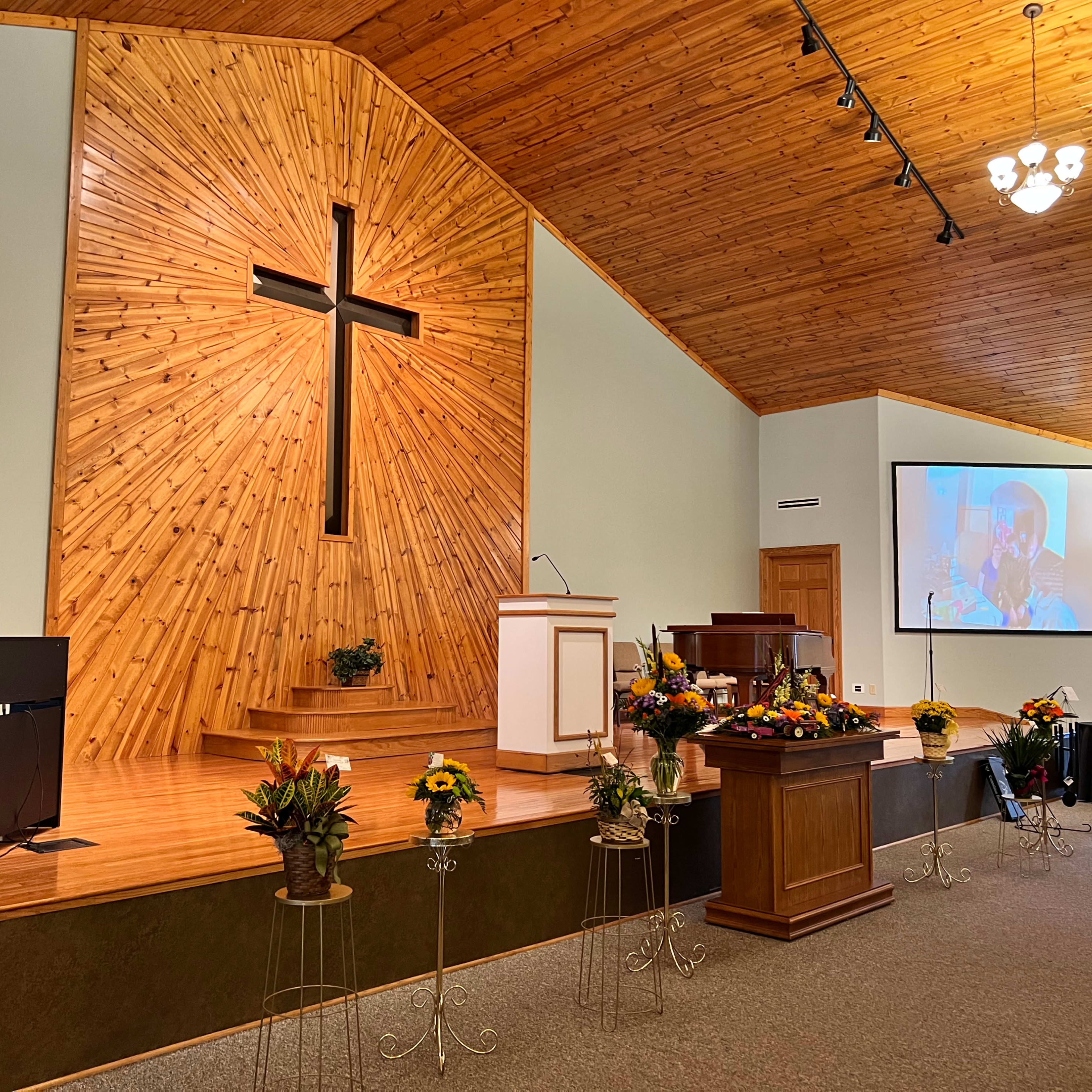 An interior view of a church with a wooden cross on the wall and a stage area featuring floral arrangements and a large screen displaying a person.